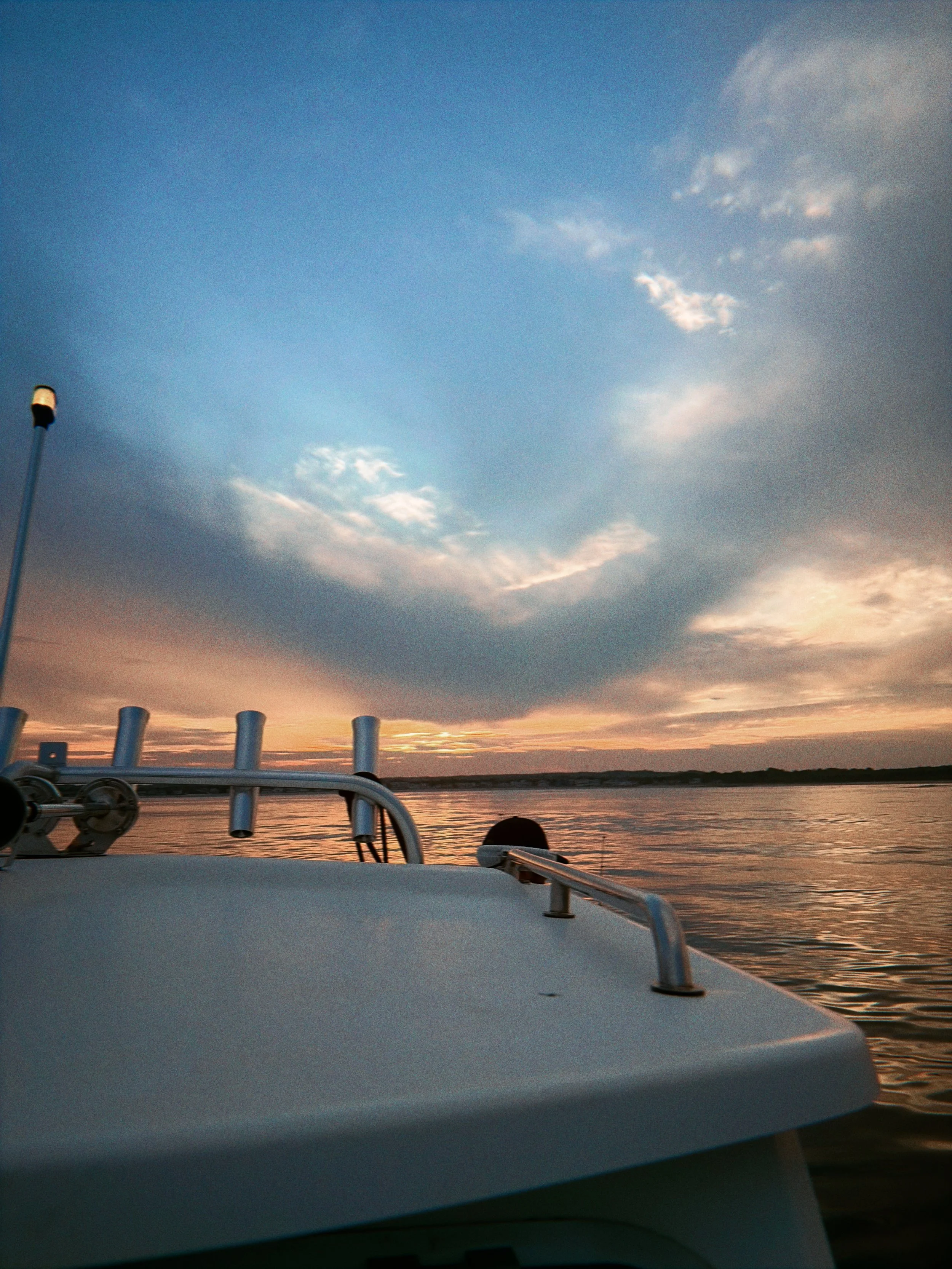 View from a boat at sunset or sunrise with a cloudy sky and water in the background, a pole on the left, and part of the boat's railing in the foreground.