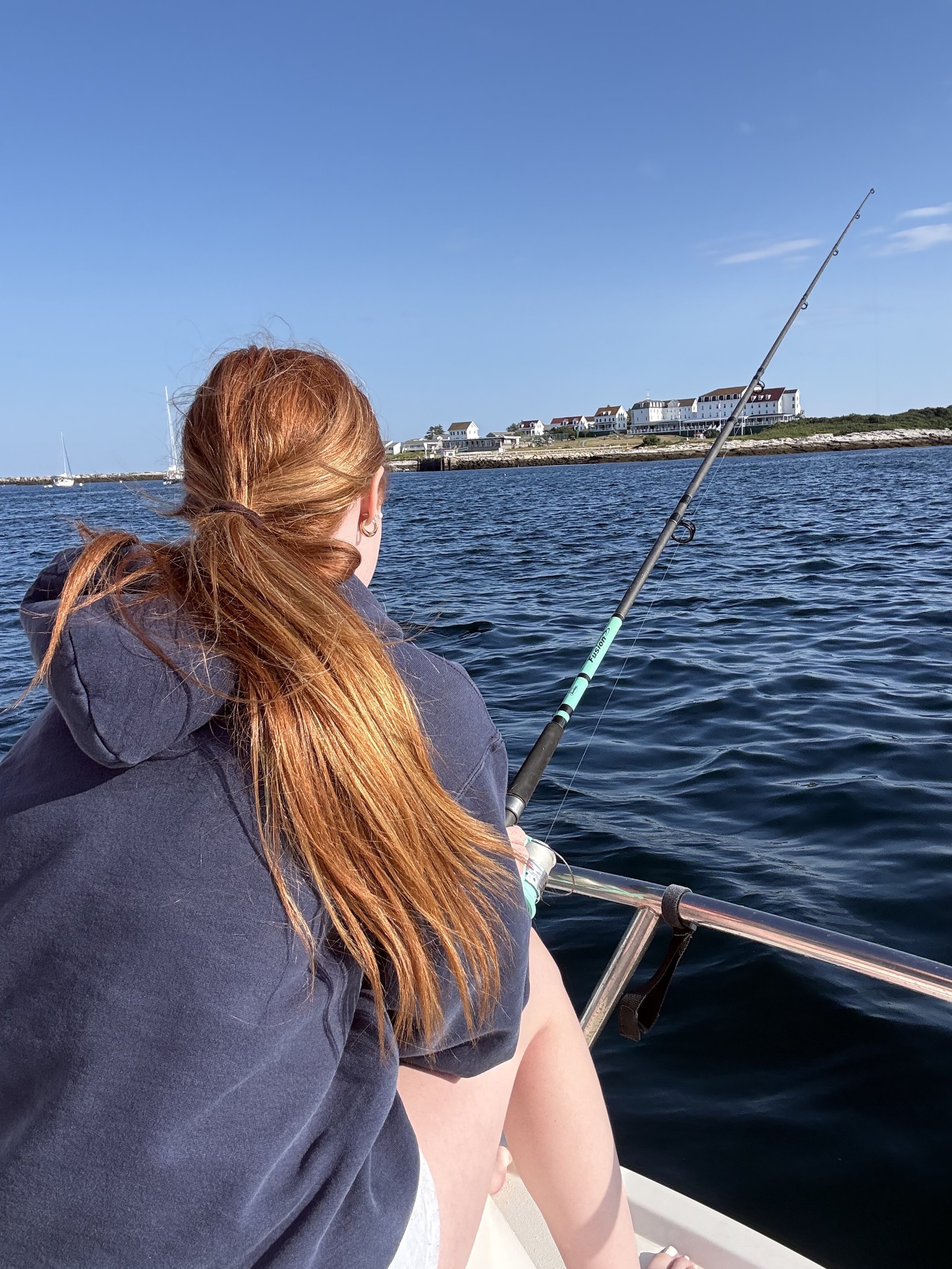 A woman with long red hair fishing off a boat onto a body of water, with buildings on the shoreline and clear blue sky in the background.