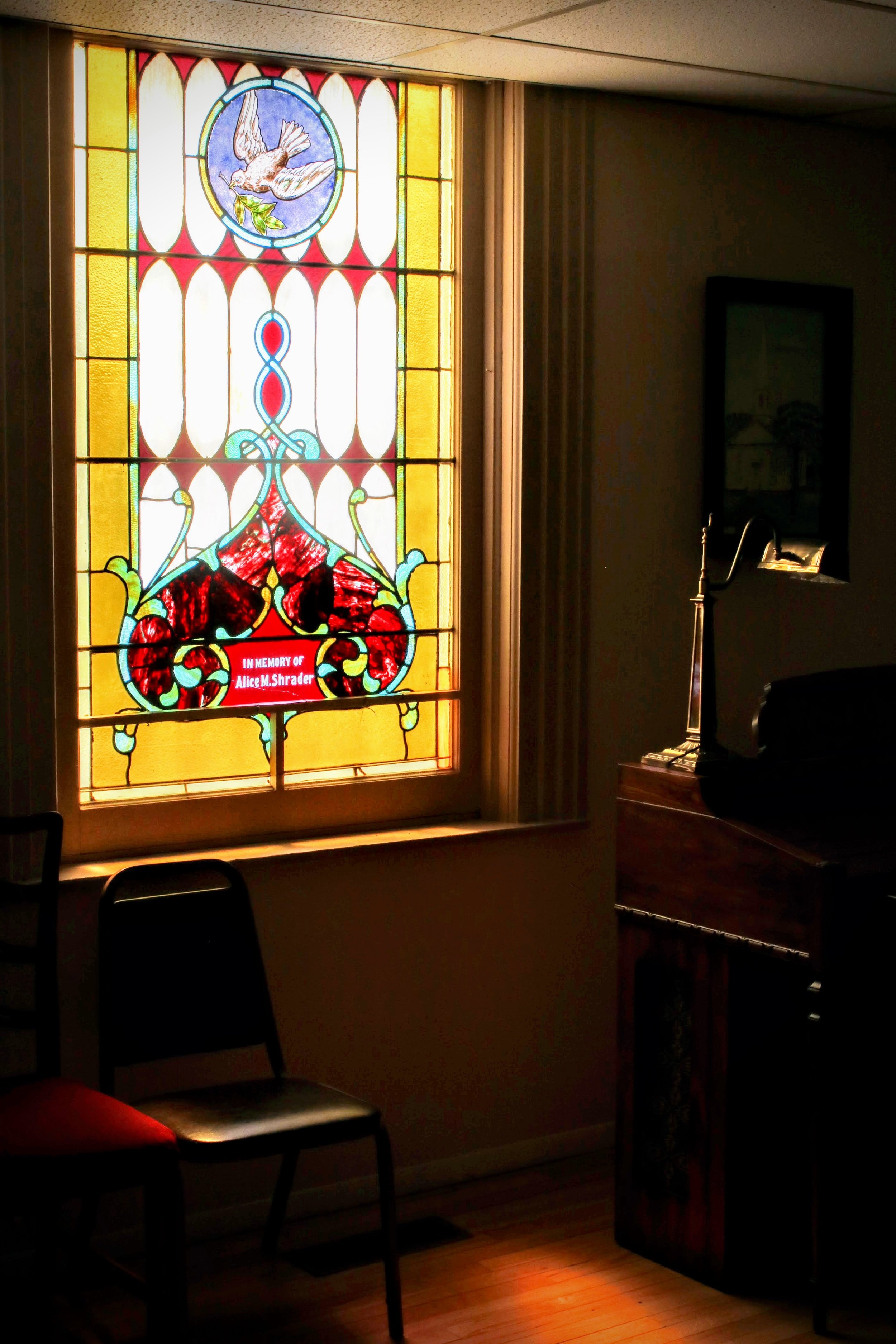 A stained glass window depicting a dove holding an olive branch, with yellow and red decorative borders and a red plaque in memory of Alice M. Shrader.