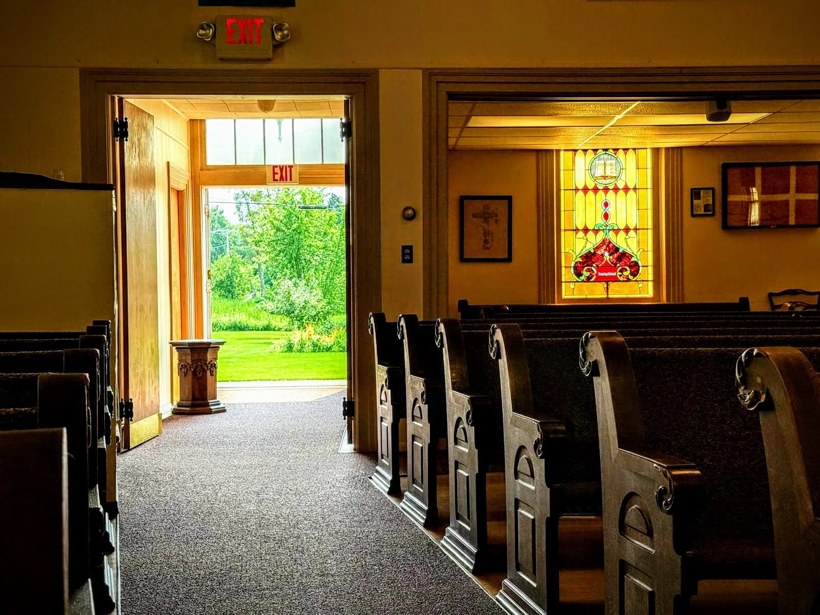 Interior of a church or chapel with wooden pews, stained glass window, open door to outside, and greenery visible through the door.