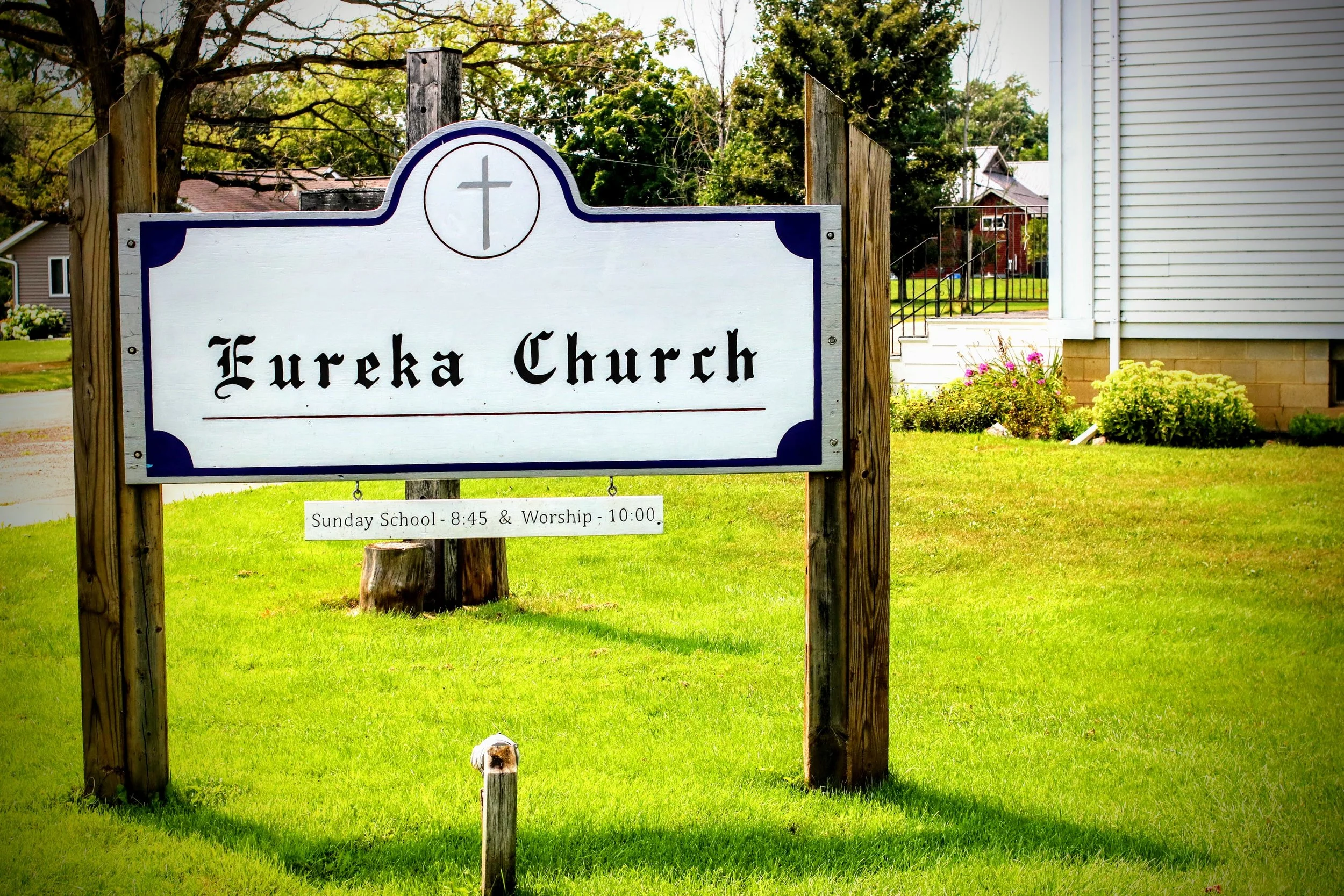 Sign outside Eureka Church with a cross symbol at the top and text indicating Sunday school at 8:45 and worship service at 10:00, surrounded by green grass, plants, and a white building in the background.