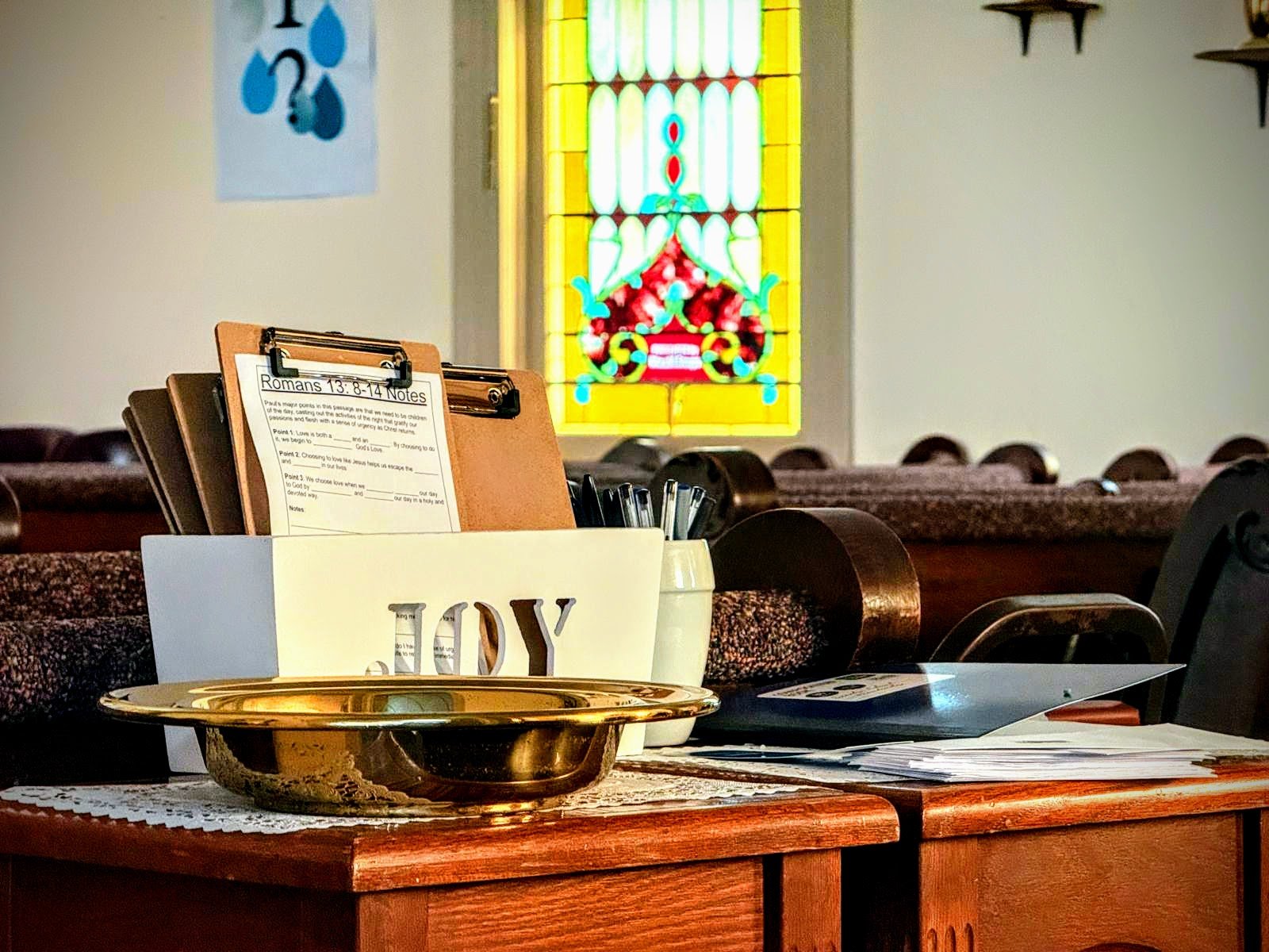 Inside a church, a table with a gold dish, a white mug with the word 'DAY' visible, papers, and a small container holding pens. In the background, a colorful stained glass window and a blue poster with a question mark and water drops on the wall.