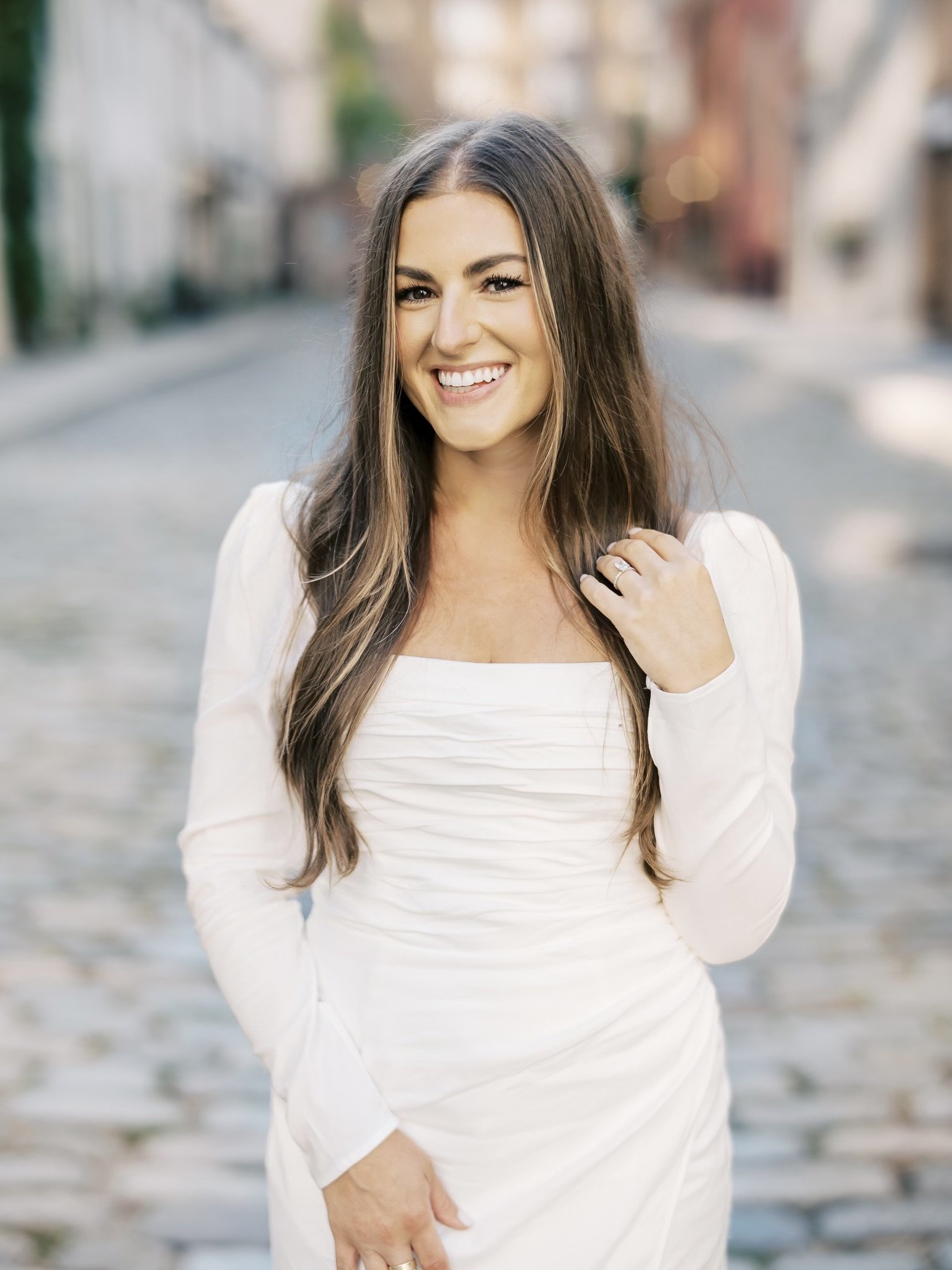 A woman in a white dress smiling outdoors, standing near a body of water with buildings and trees blurred in the background.