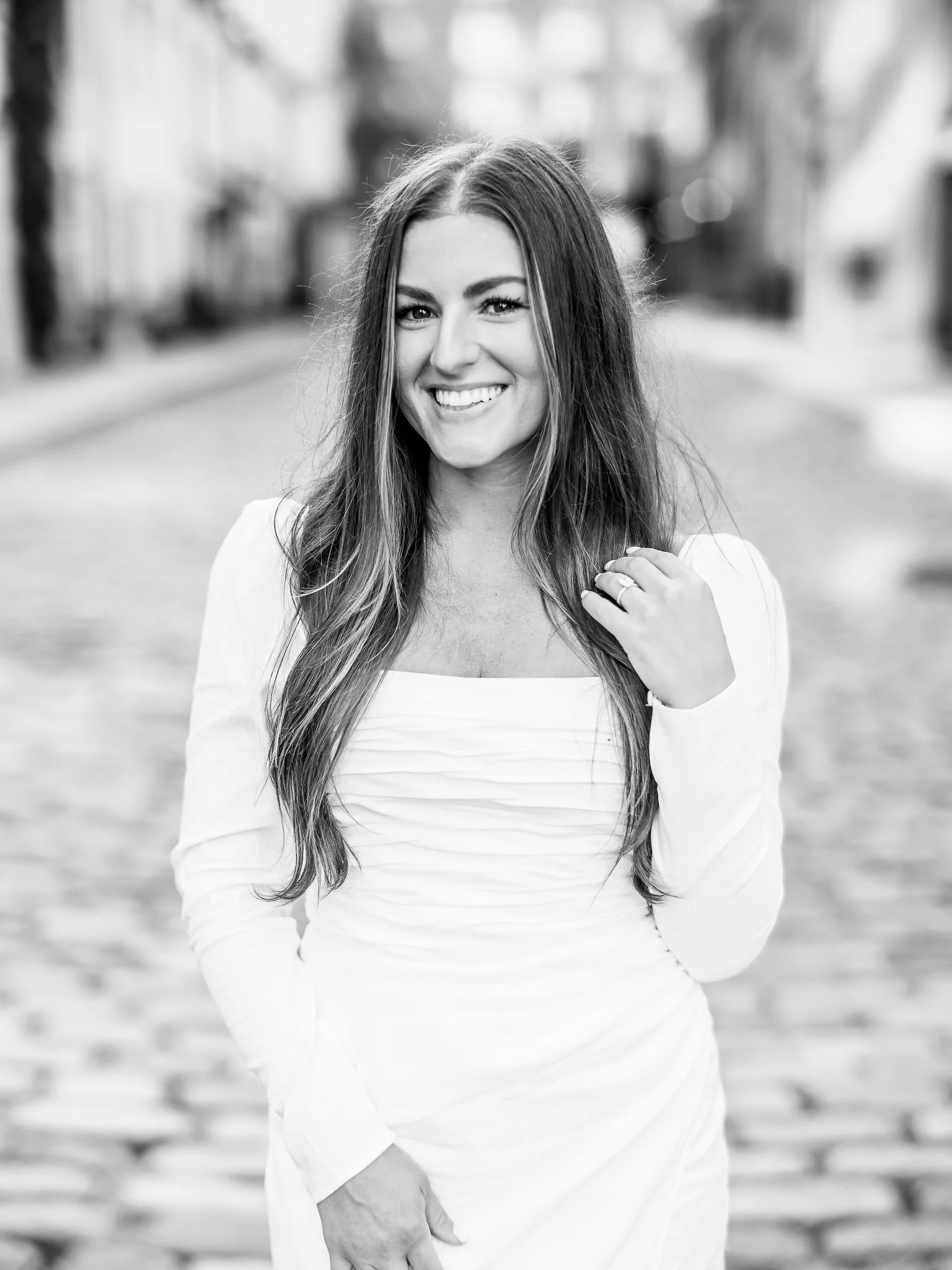 Black and white photo of a smiling young woman with long hair, standing outdoors with a blurred background of trees and a bench.