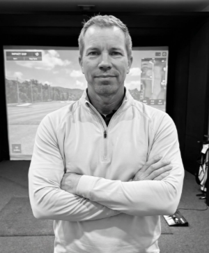 A man standing with arms crossed inside an indoor golf simulation room with a screen showing a golf course behind him.