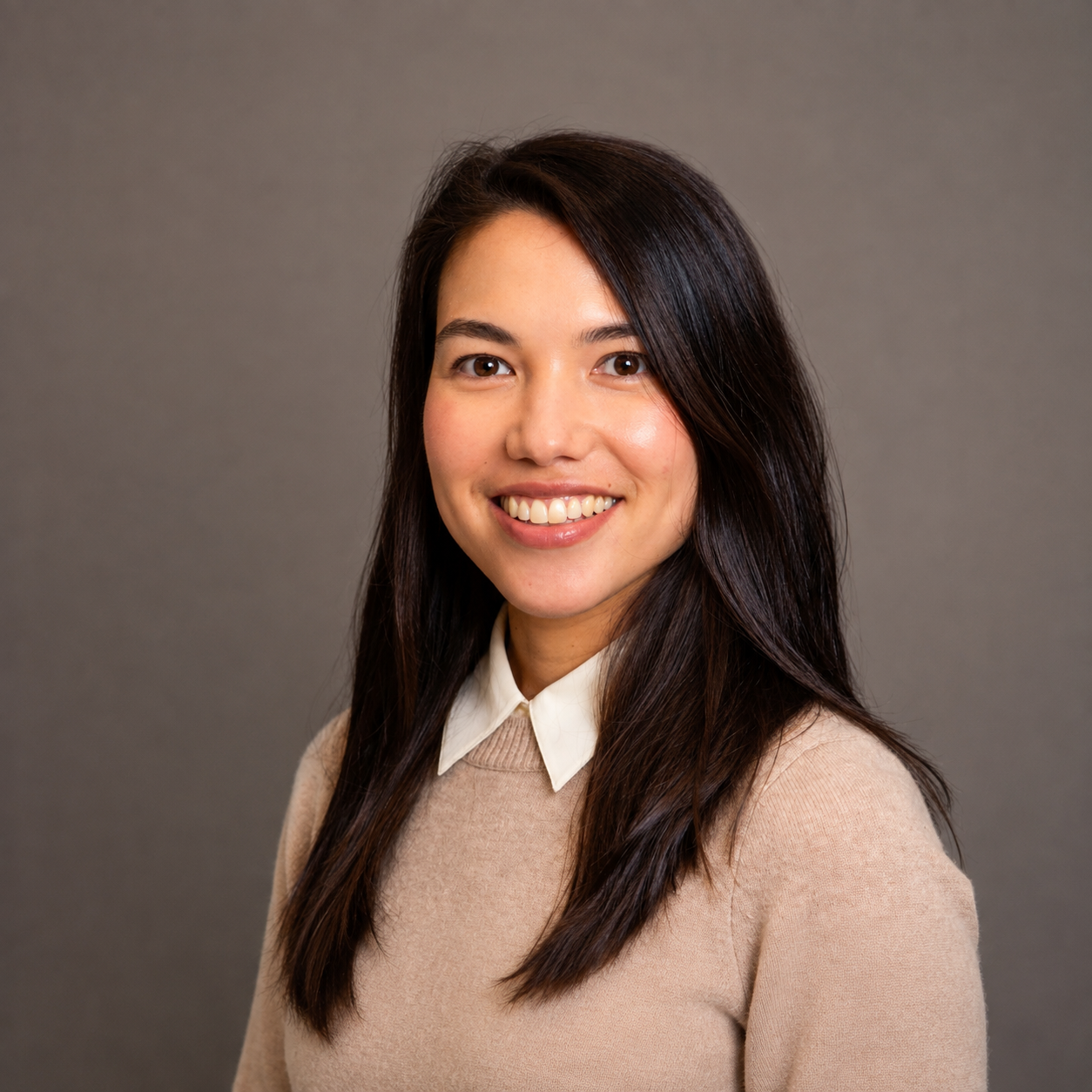 A portrait of a young woman with long dark brown hair, smiling, wearing a beige sweater over a white collared shirt, against a plain gray background.