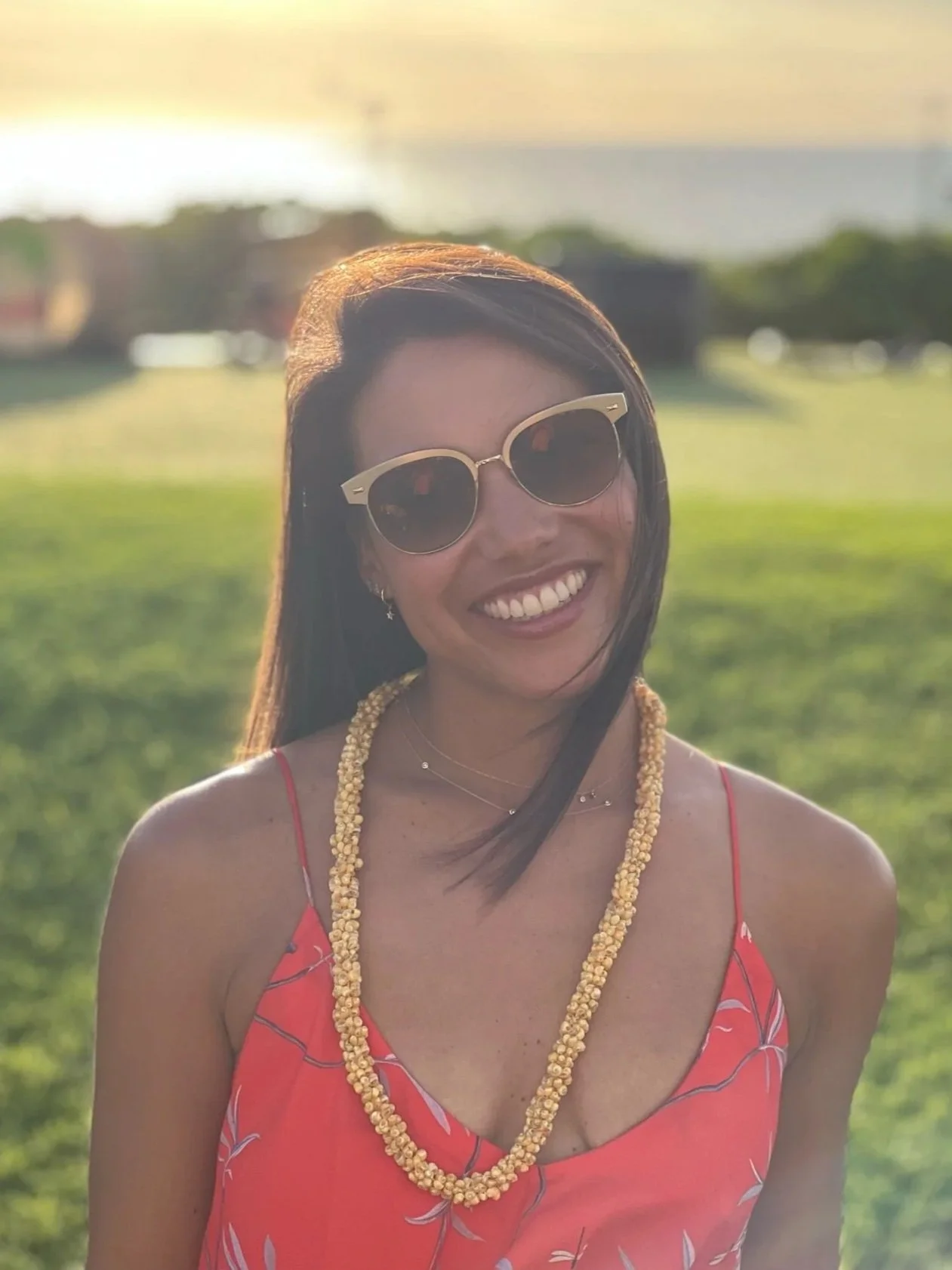 Smiling woman wearing sunglasses, a coral dress with a leaf pattern, and a beaded necklace in an outdoor setting during sunset
