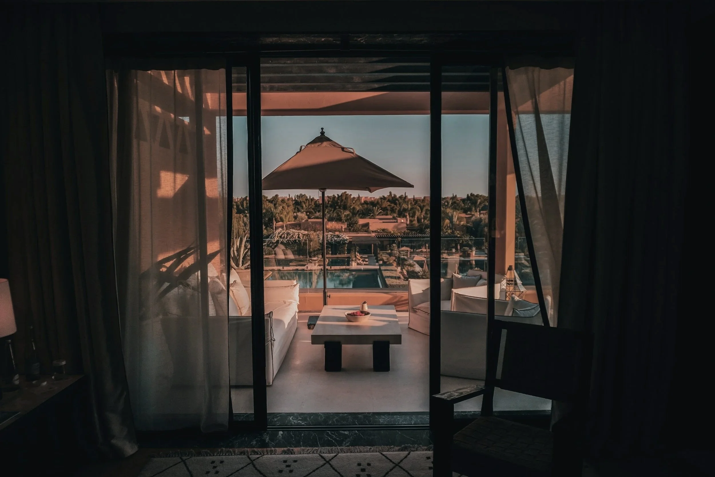 Interior view of a room with large glass doors leading to a balcony with outdoor furniture and a sunshade, overlooking a landscape with trees and buildings, during sunset.