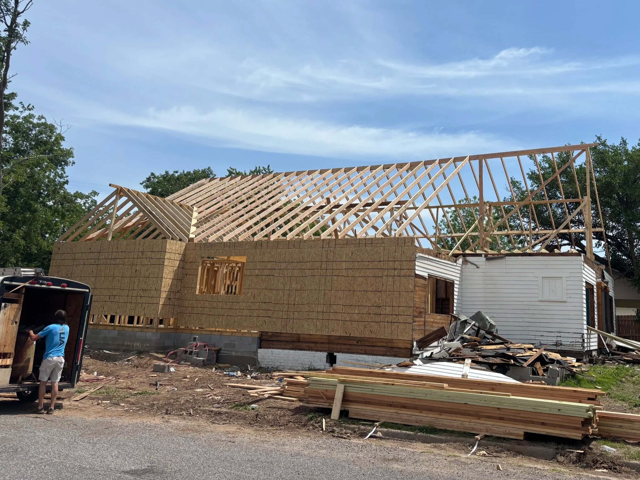 A house under construction with a wooden framework for the roof, surrounded by trees and construction debris.