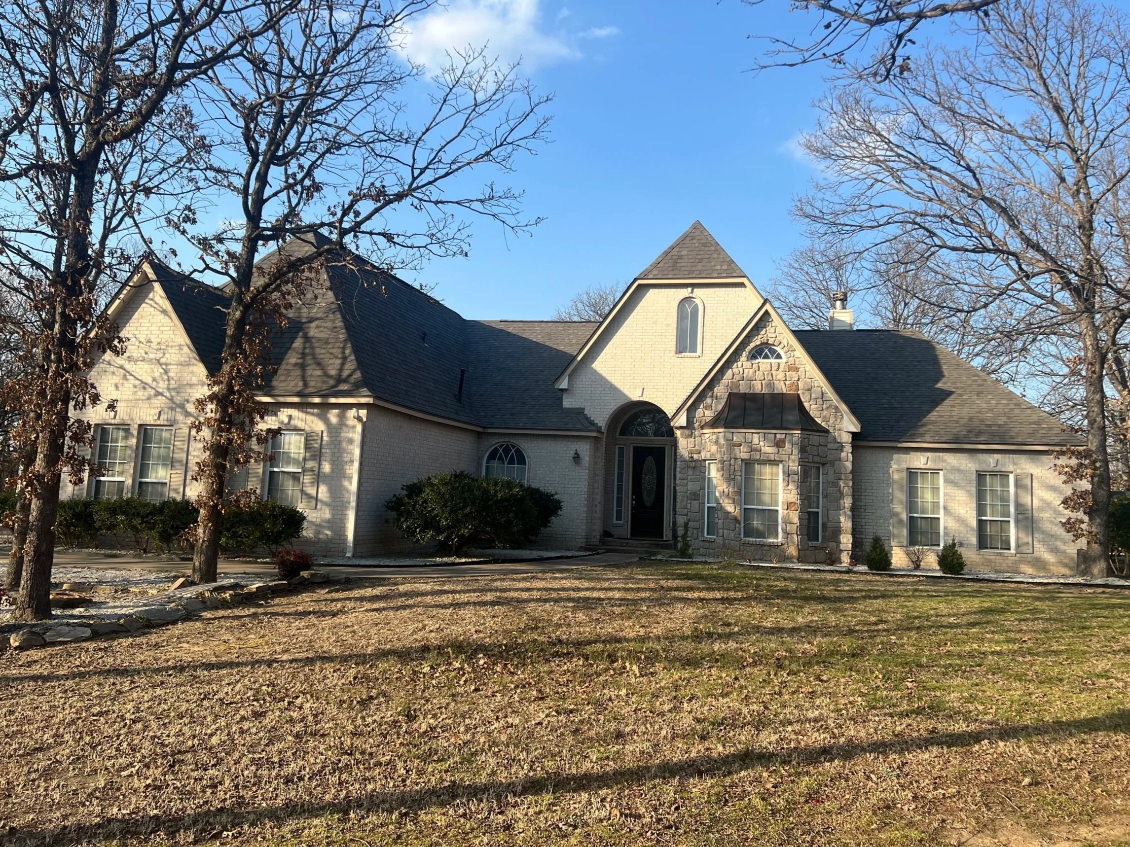 A two-story house with a combination of white brick and stone exterior, multiple gables, and large windows. The house is surrounded by leafless trees and a front yard with patches of grass and dirt.