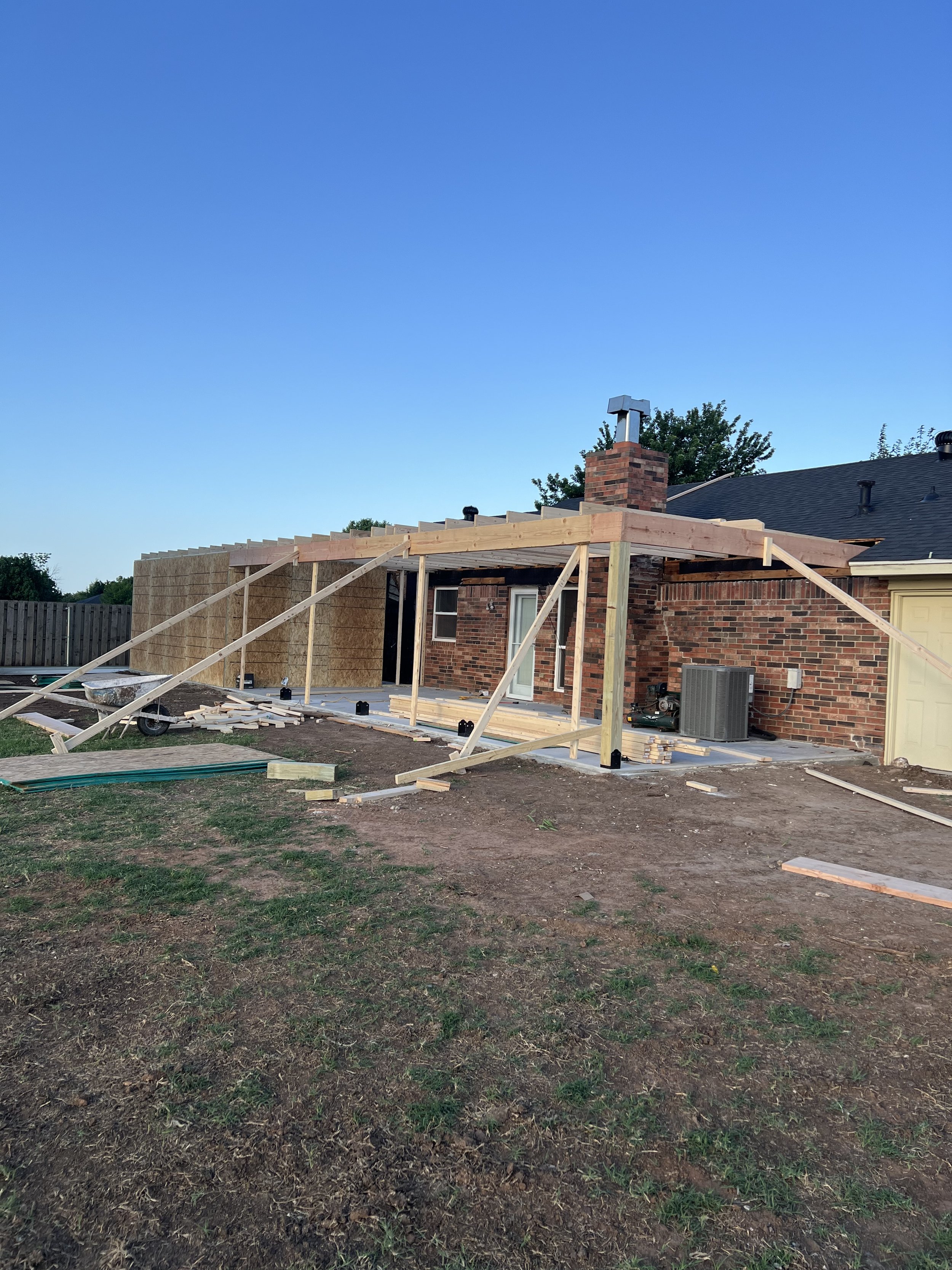 Construction work on the backyard of a brick house with a new wooden structure being built. There is a chimney on the house, a window and an air conditioning unit visible. The ground is partially grass and dirt. The sky is clear and blue.