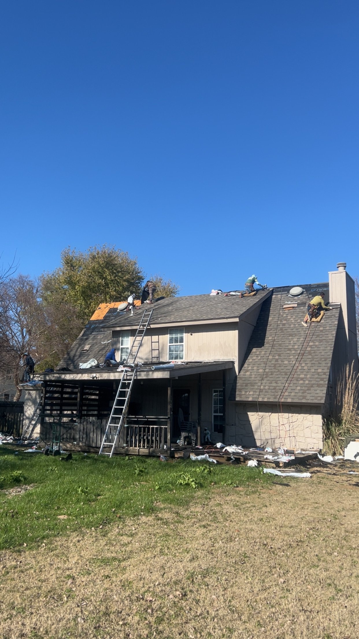 People working on a roof of a house on a sunny day, with ladders and roofing materials visible, and a grassy yard in front.