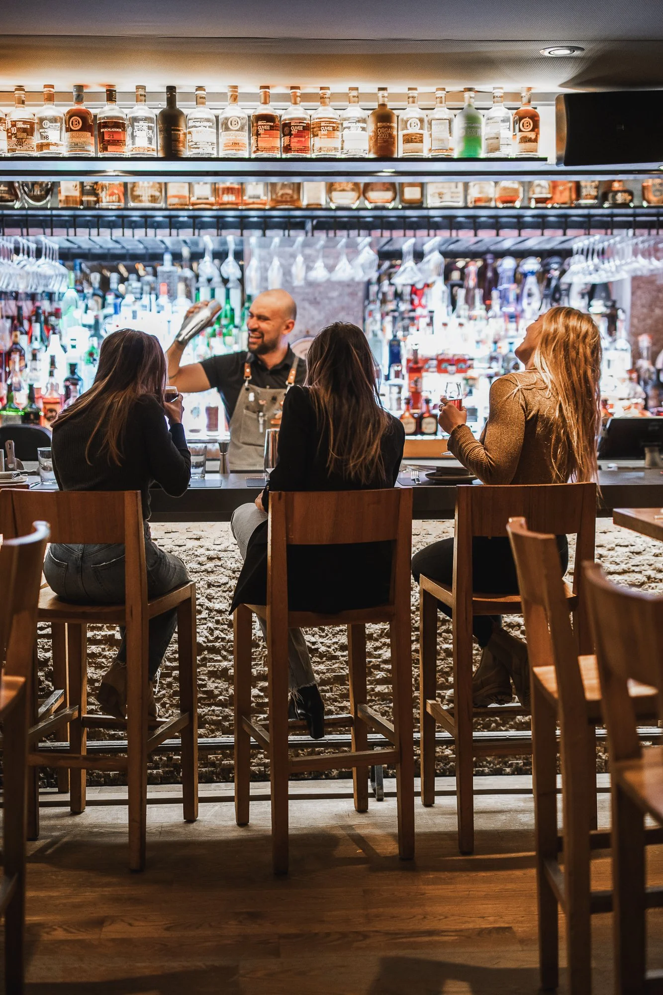 Bar scene with three women seated at the counter, bartender pouring a drink, bottles of alcohol on shelves in the background.