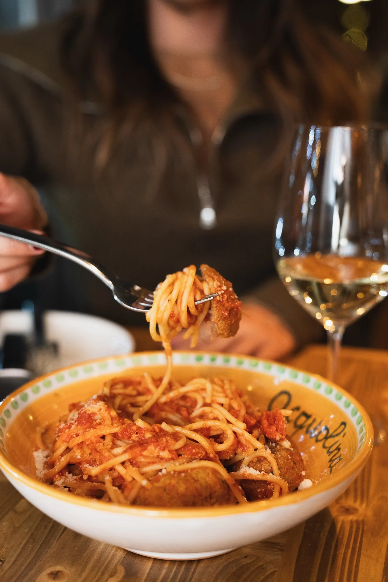 A bowl of spaghetti with meatballs in marinara sauce, topped with cheese. A fork is lifting some noodles from the bowl. There is a glass of white wine beside the bowl.