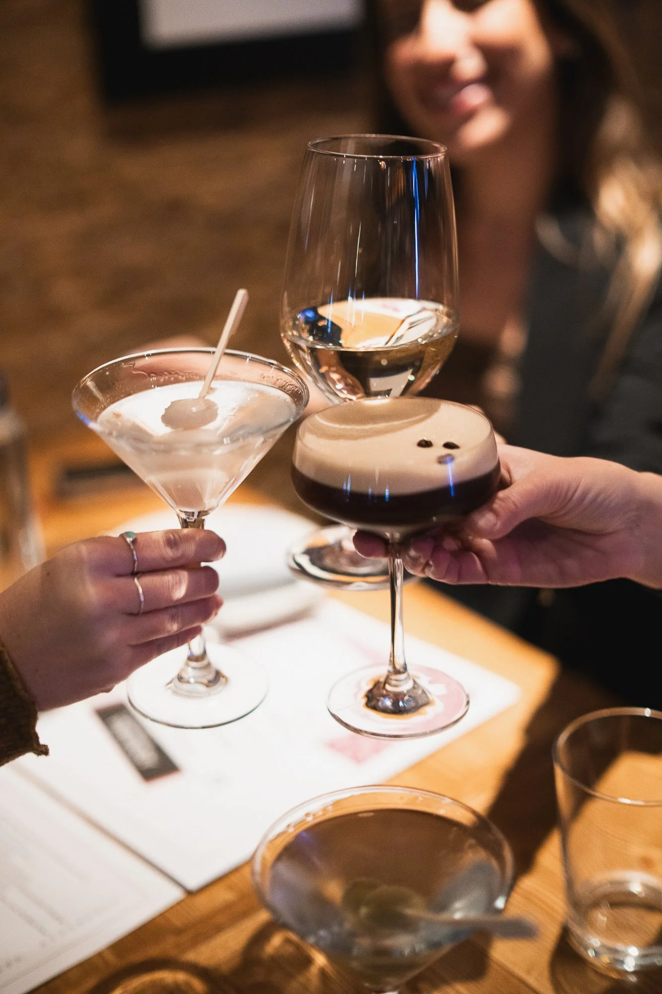 People toasting with various glasses of drinks including a martini, white wine, and a dark cocktail with foam, in a dimly lit setting.
