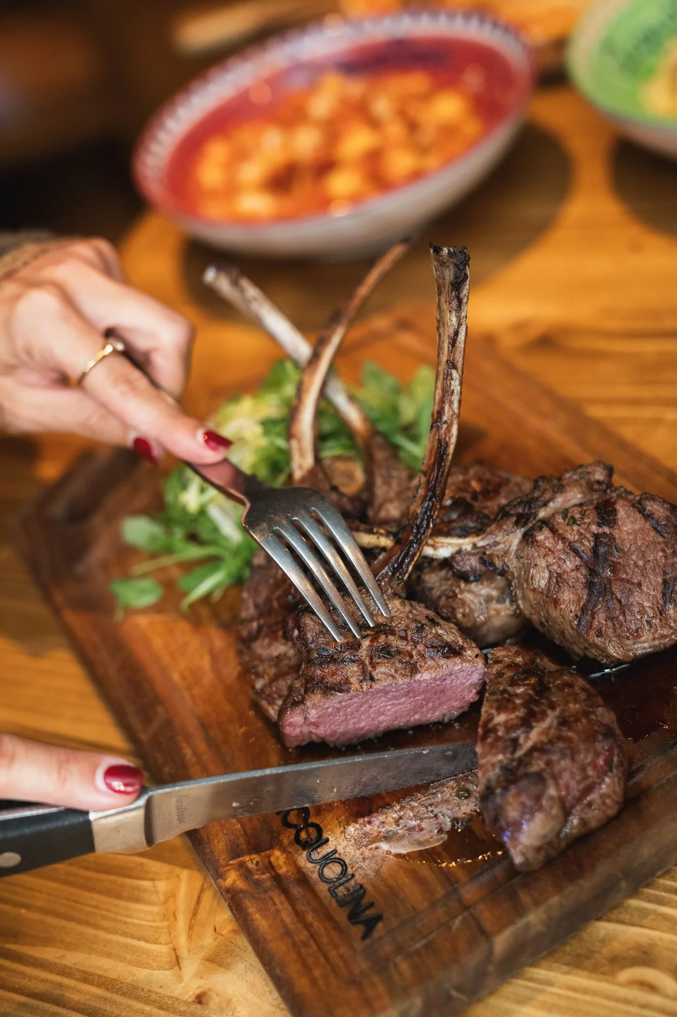 Person cutting into a cooked rack of lamb, with a bowl of sauce and other dishes blurred in the background on a wooden table.