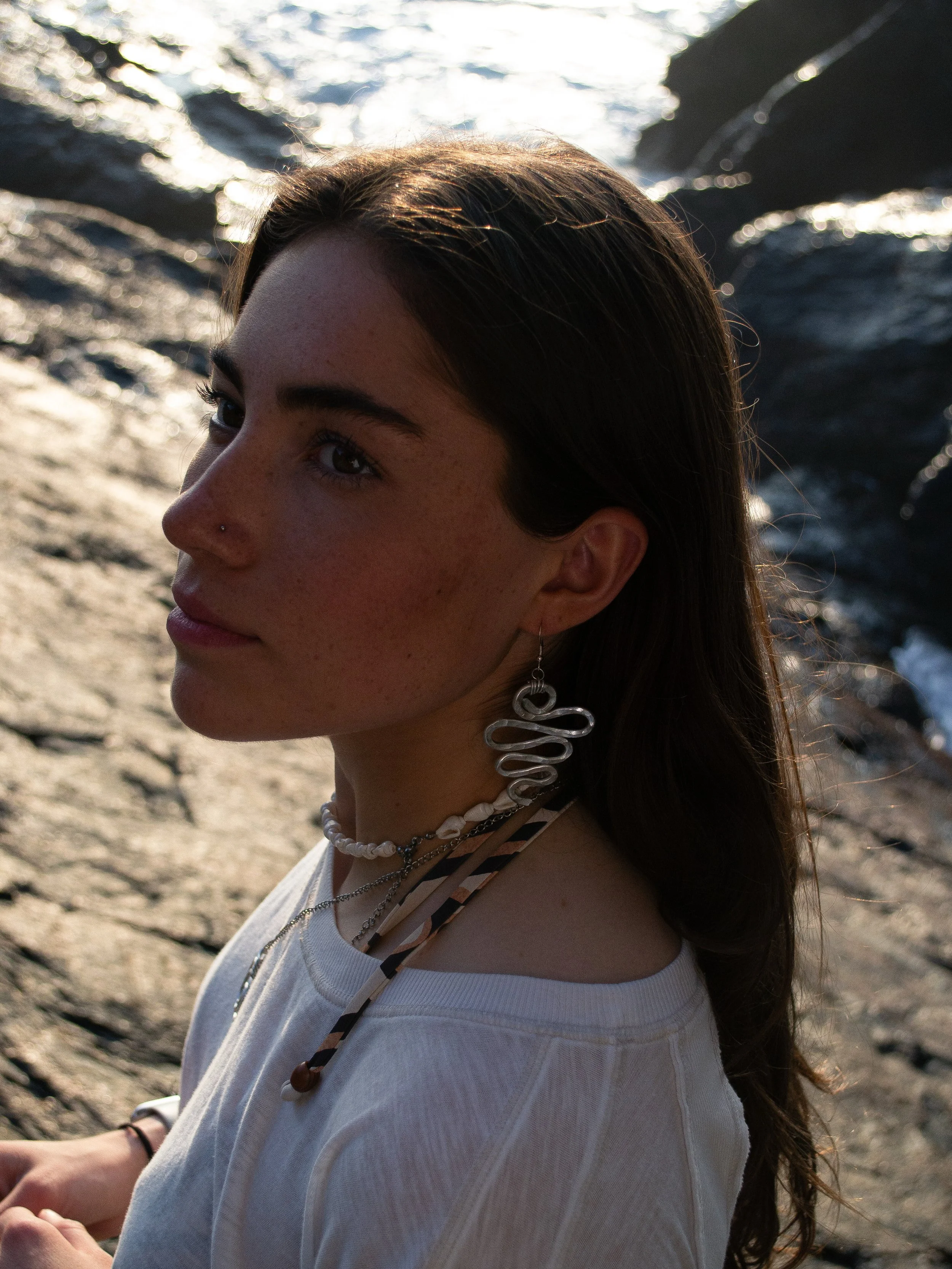 A young woman with long brown hair and snake earrings sitting on a rocky beach near the water at sunset, looking away.