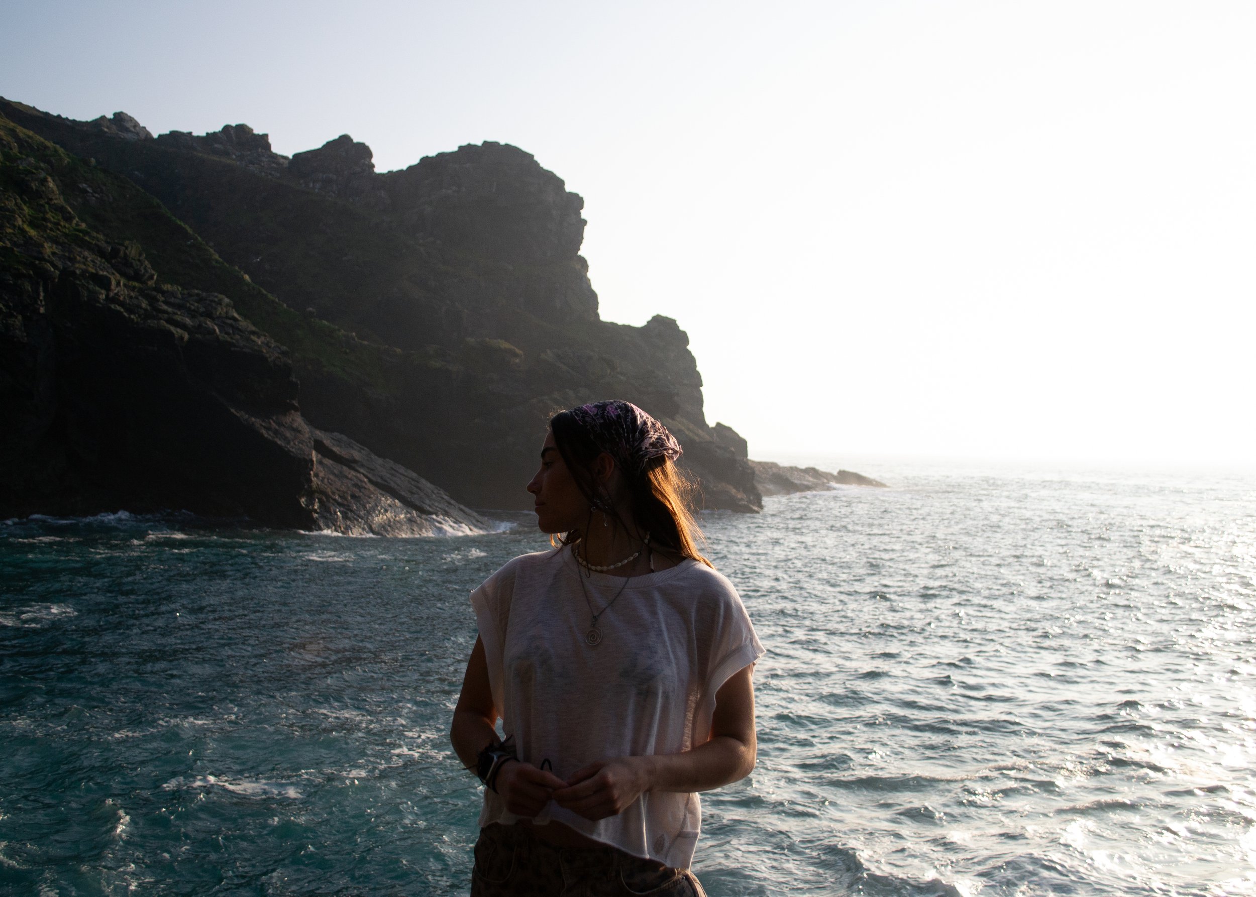 A woman standing by the ocean on a rocky coastline during sunset, with cliffs in the background wearing the spiral necklace.