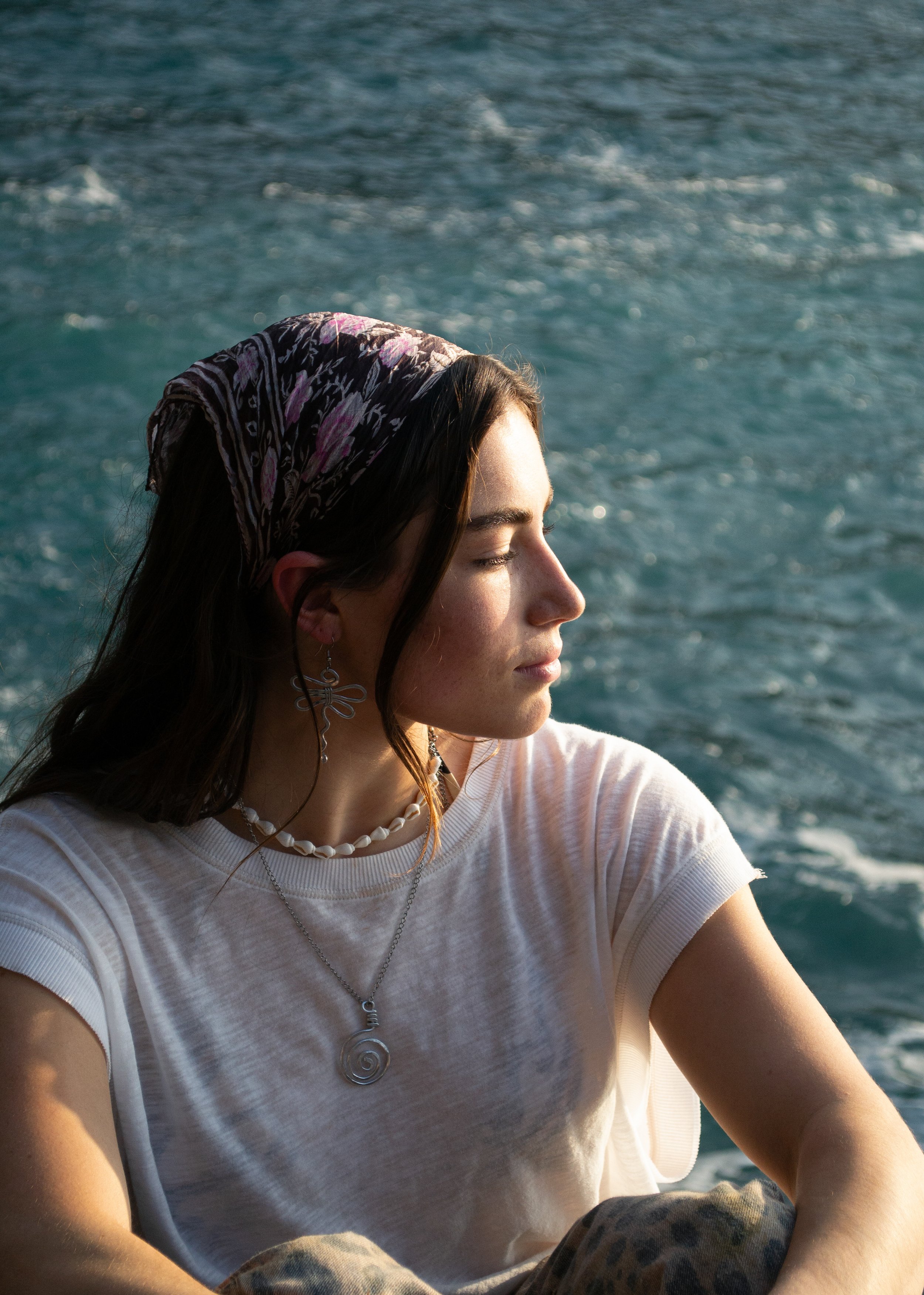 A young woman with closed eyes and long brown hair, wearing by hammered dragonfly earrings, a spiral necklace, sitting by the water with a blurred ocean background.