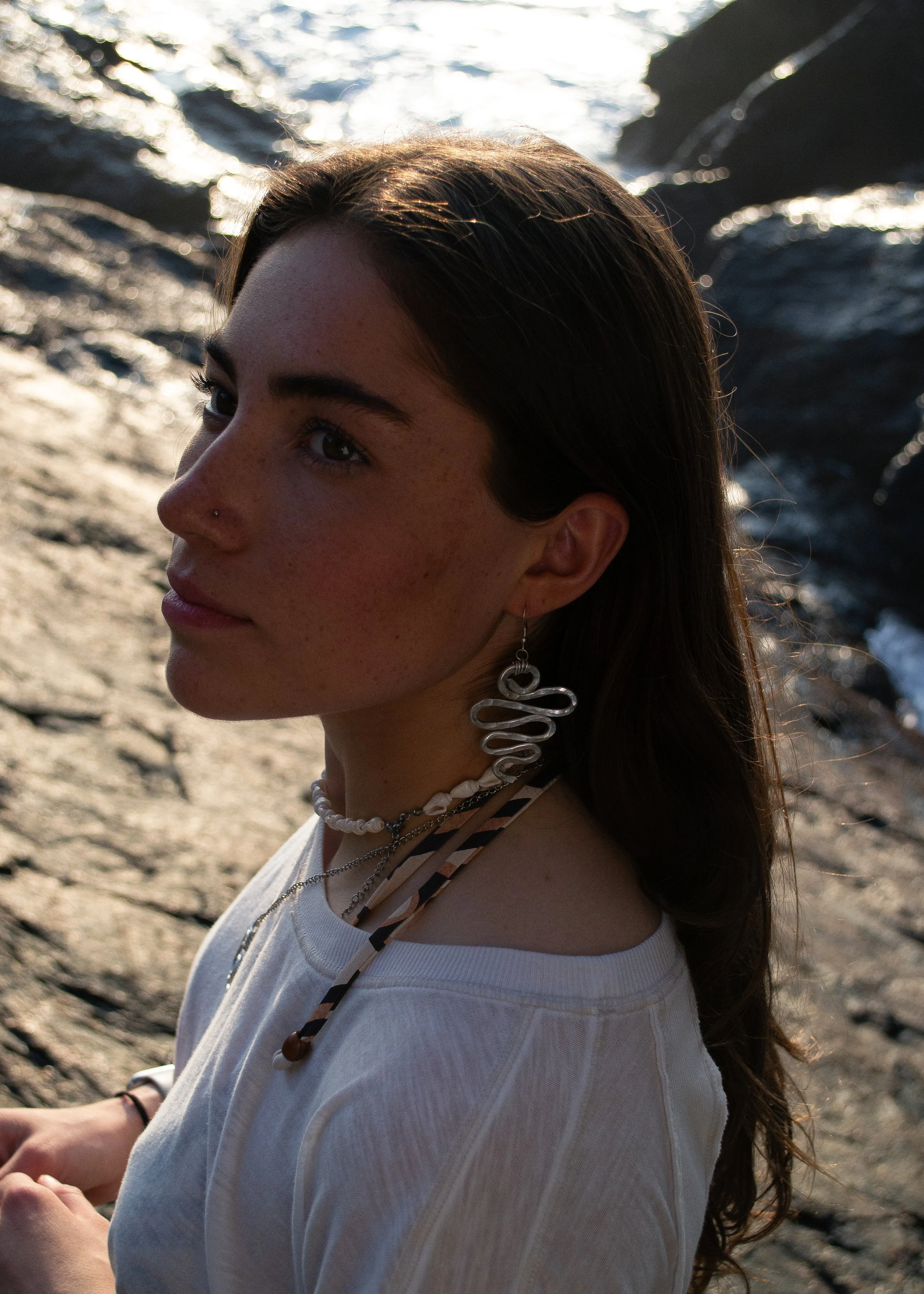 A young woman with brown hair, wearing multiple necklaces and earrings, appears to be sitting on a rocky shoreline with water in the background, illuminated by sunlight.