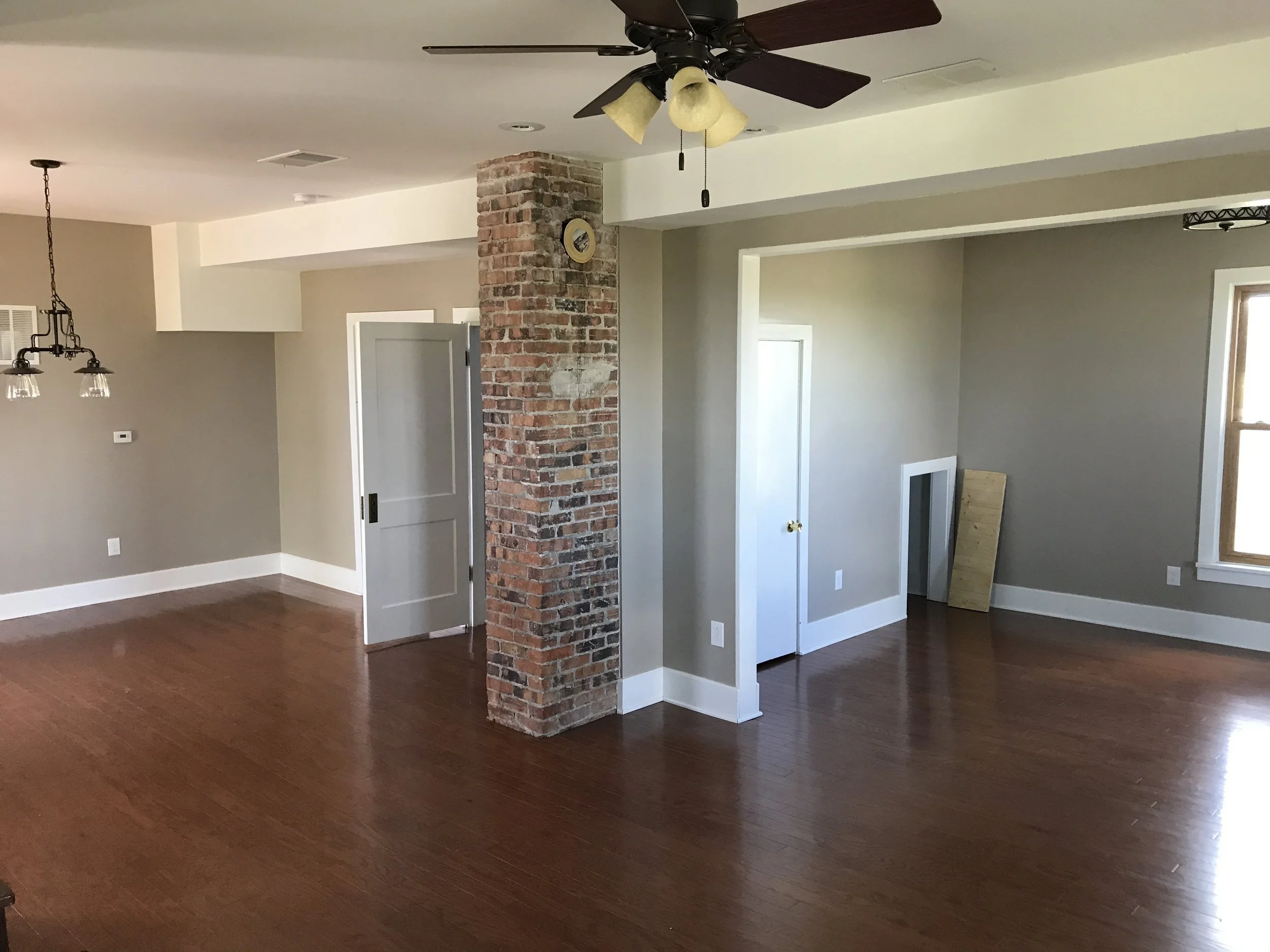 Living room with hardwood floors, exposed brick column, ceiling fan with lights, and multiple windows letting in natural light.