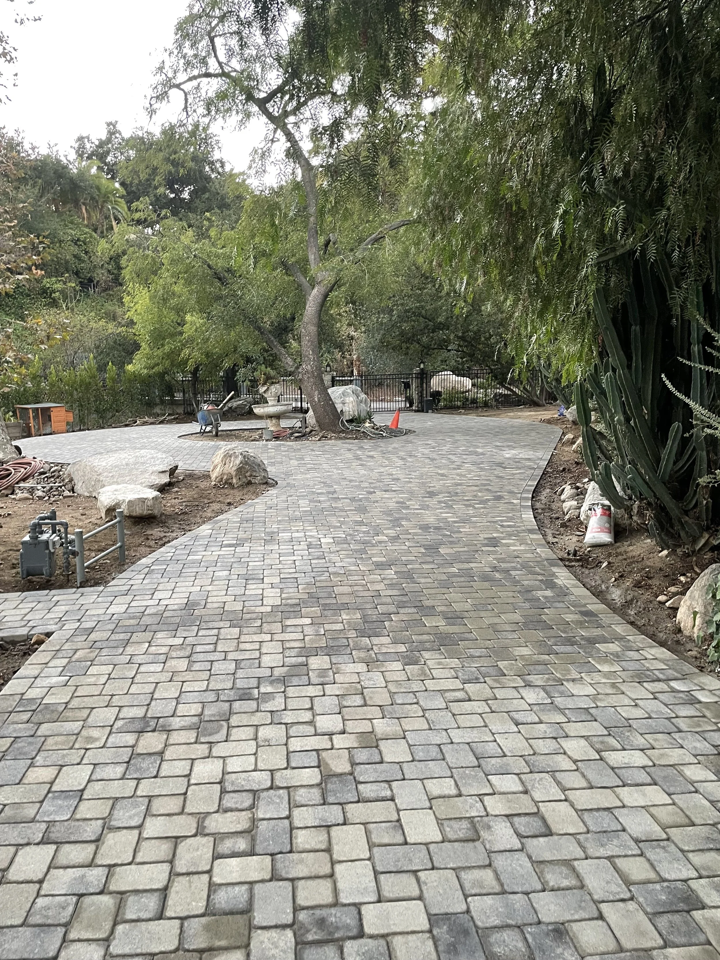 A newly paved stone pathway in a garden, surrounded by trees and plants, with construction materials and tools nearby.