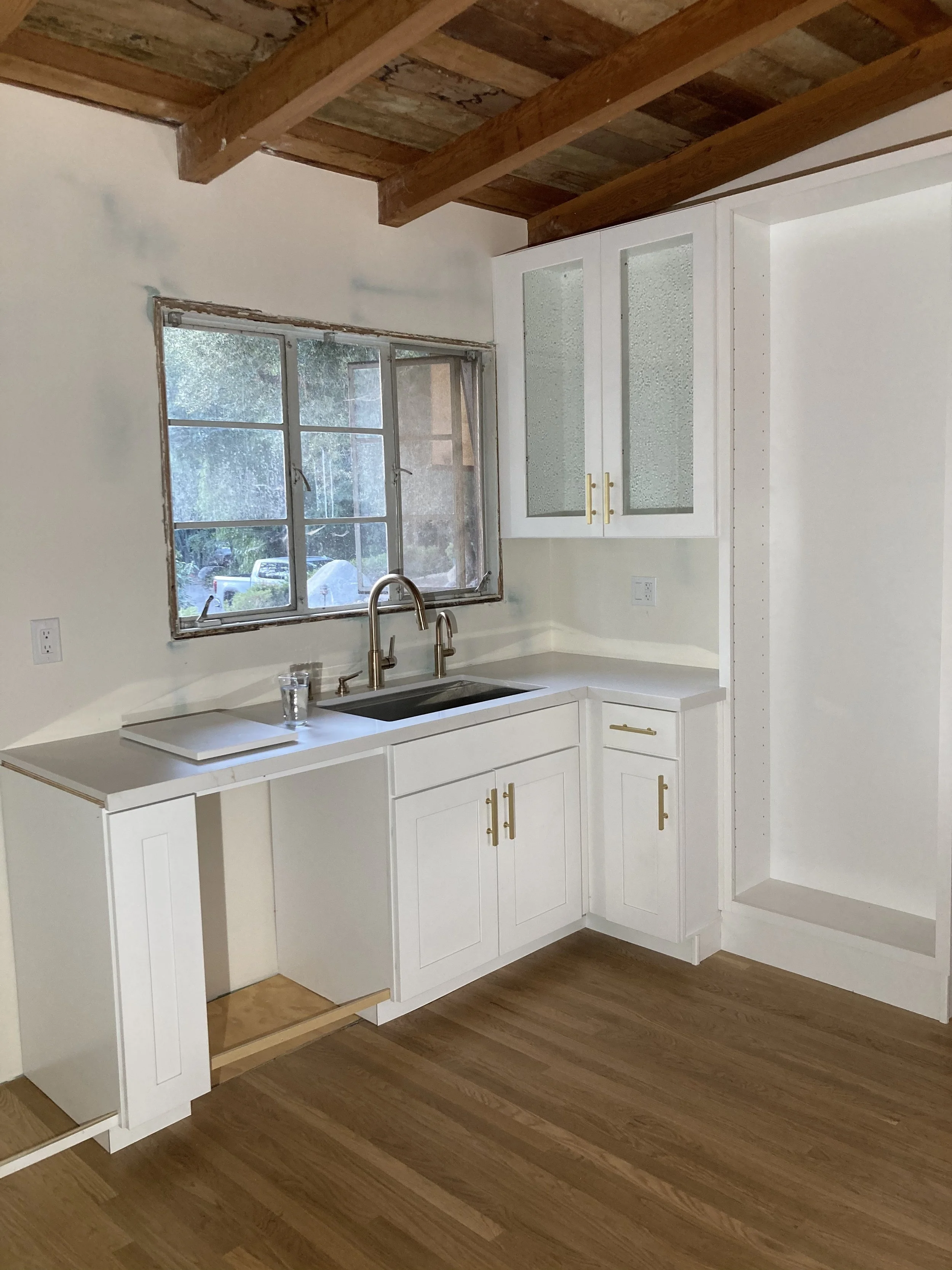A white kitchen cabinet with gold handles positioned under a window. The kitchen has wooden flooring and a wood ceiling with exposed beams.