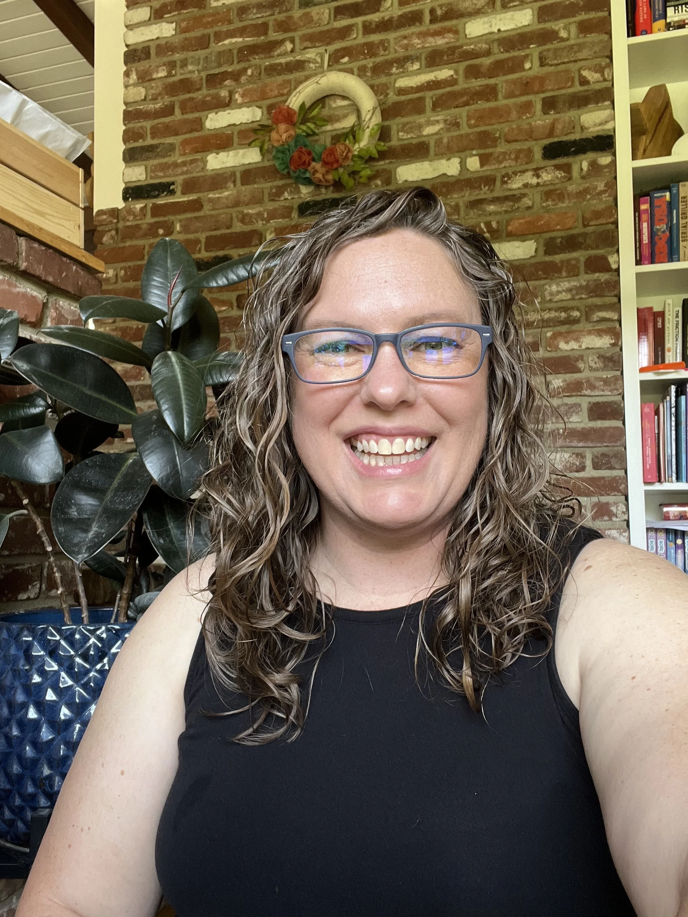 Smiling woman with curly hair and glasses indoors. Background includes a brick wall, a bookshelf, a houseplant, and decorative wall hanging.