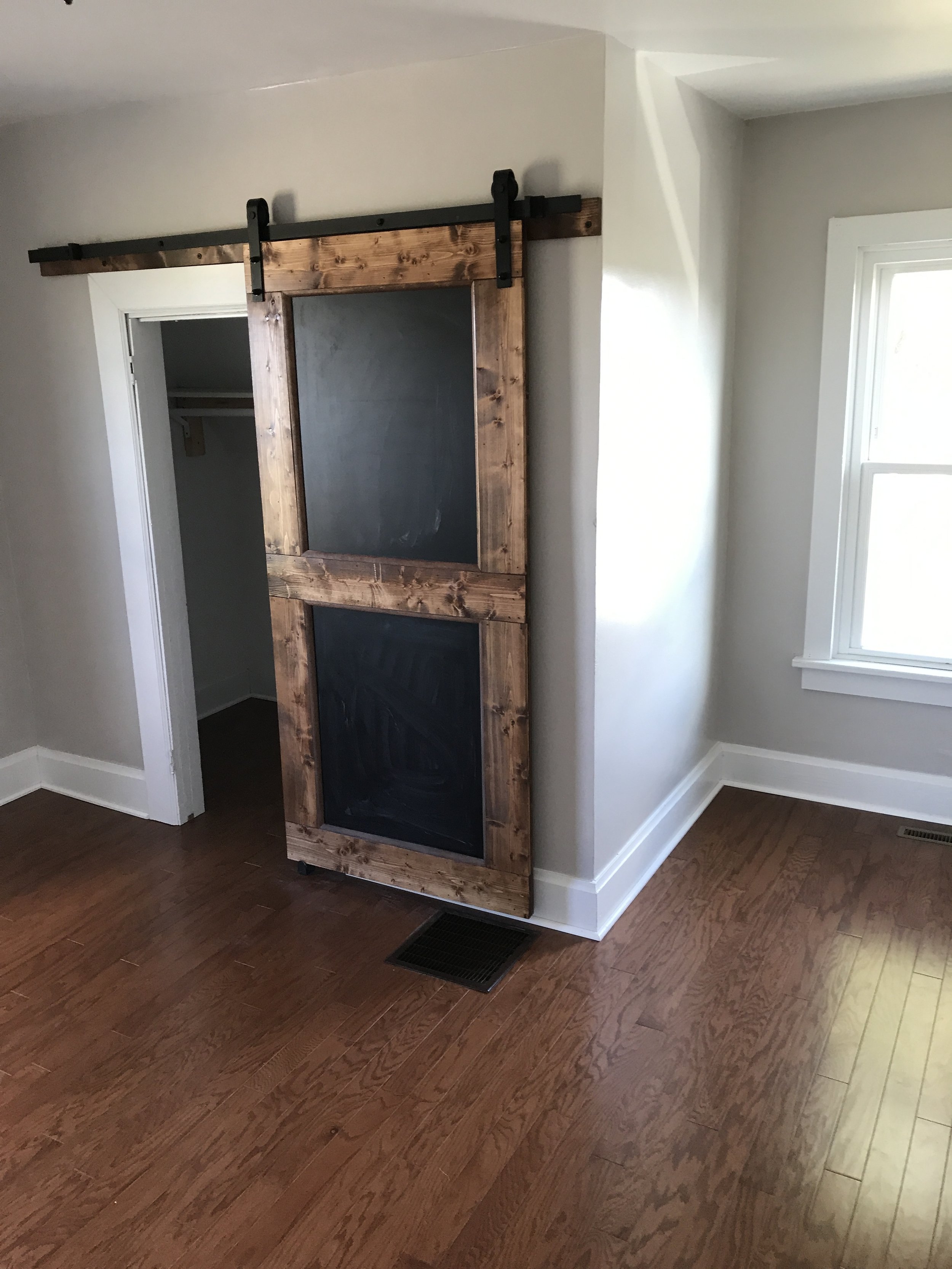 A room with hardwood floors, white walls, a window, and a sliding barn door with a chalkboard section.