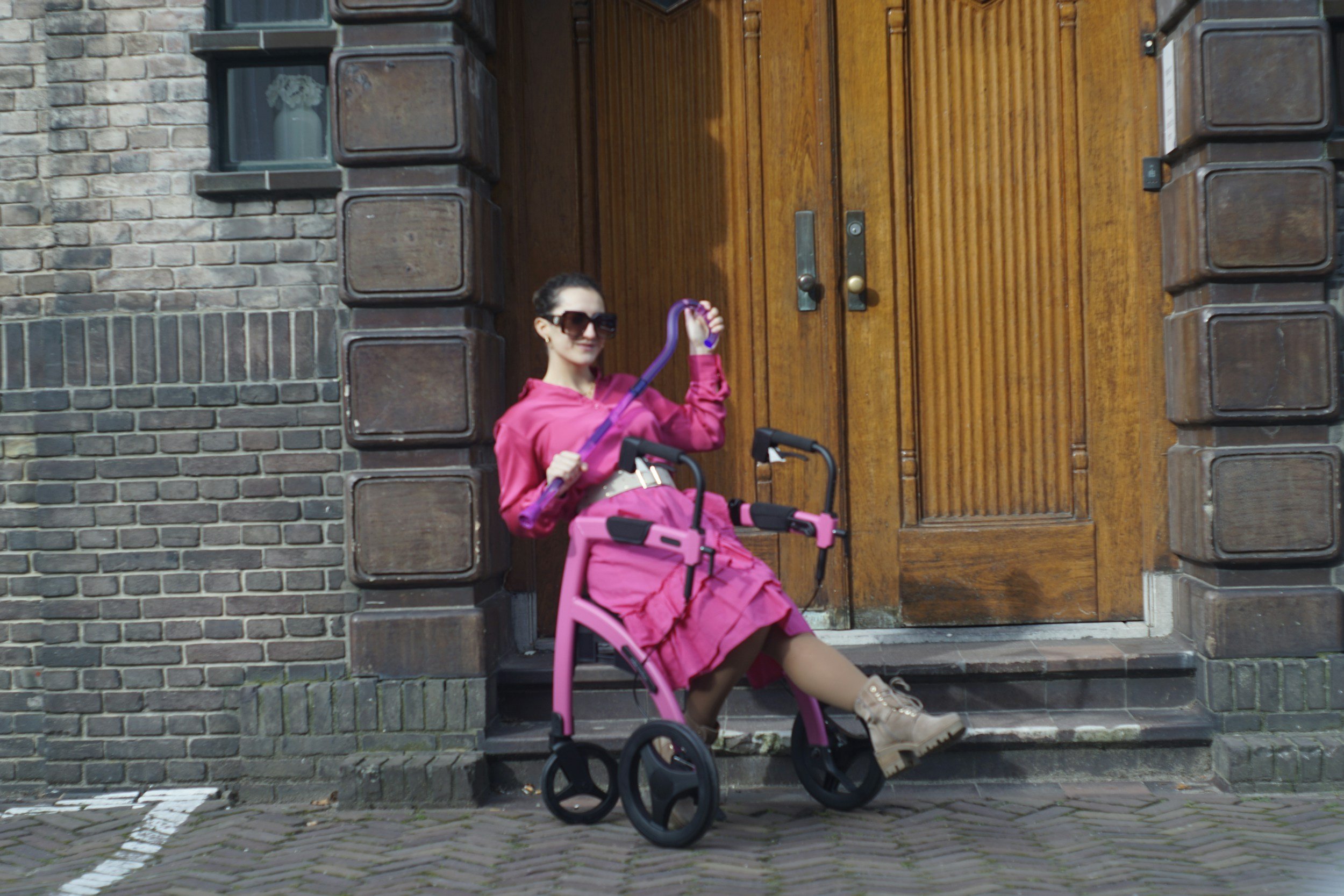A woman in a bright pink outfit and sunglasses sitting on a pink wheelchair on a steps in front of a large wooden door. She has a shower head in her hand and is smiling.