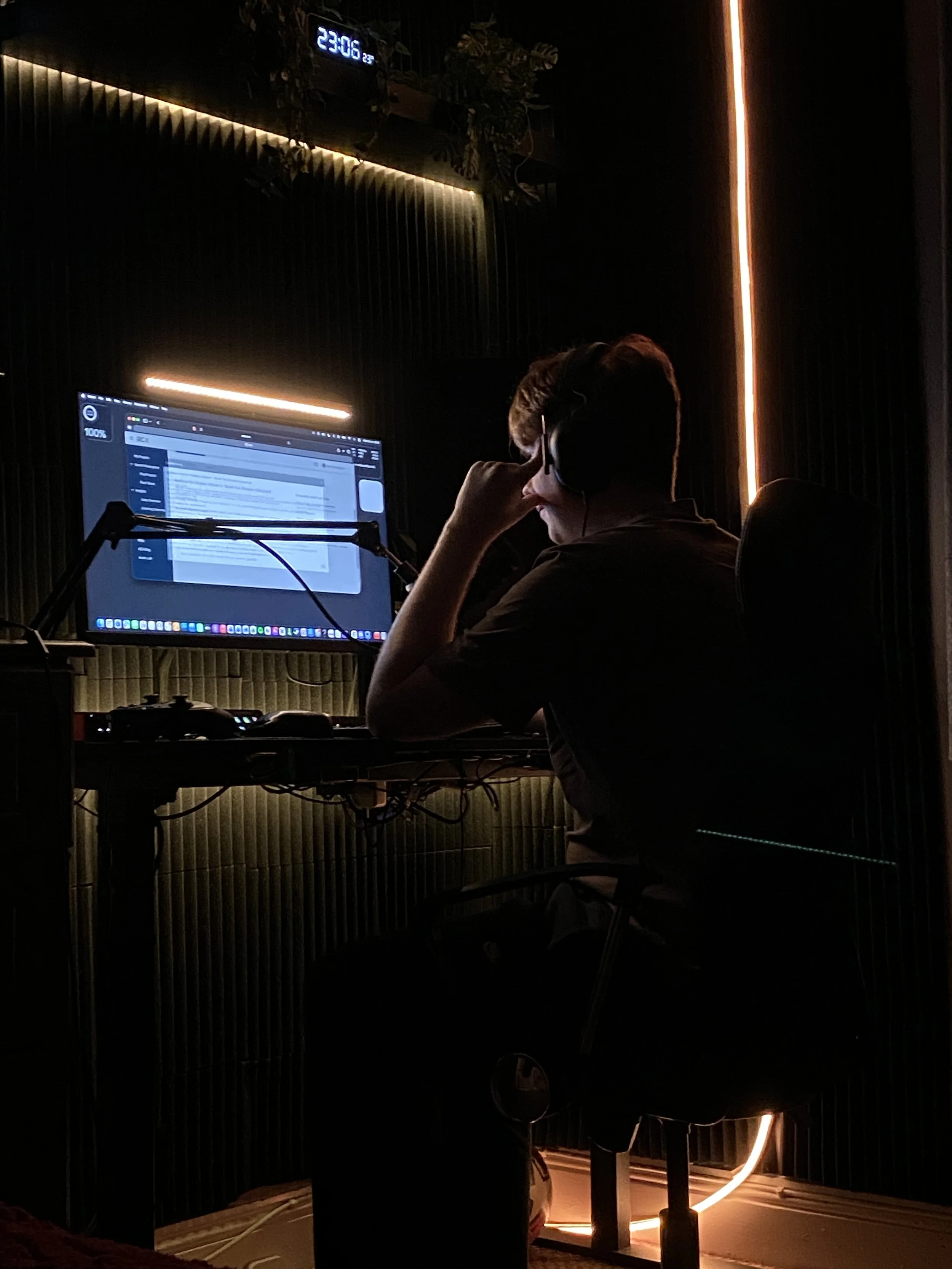 Person sitting at a desk with a computer and wearing headphones, in a dimly lit room with decorative lighting and a wall clock showing 23:06.