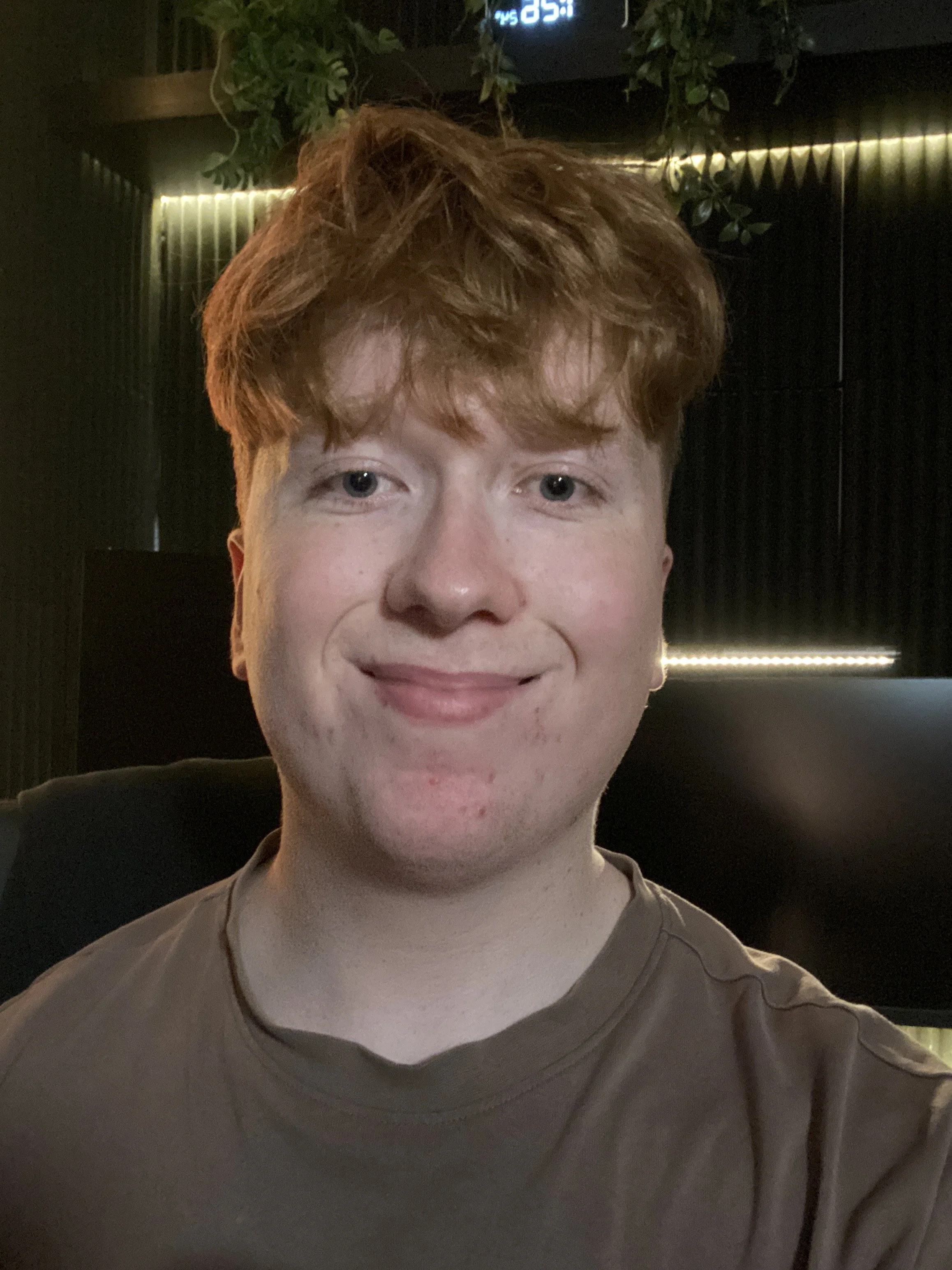 Close-up of a young man with curly red hair, light skin, and blue eyes, smiling slightly at the camera. He is wearing a tan shirt, and the background includes dark, textured walls with warm lighting and some greenery.