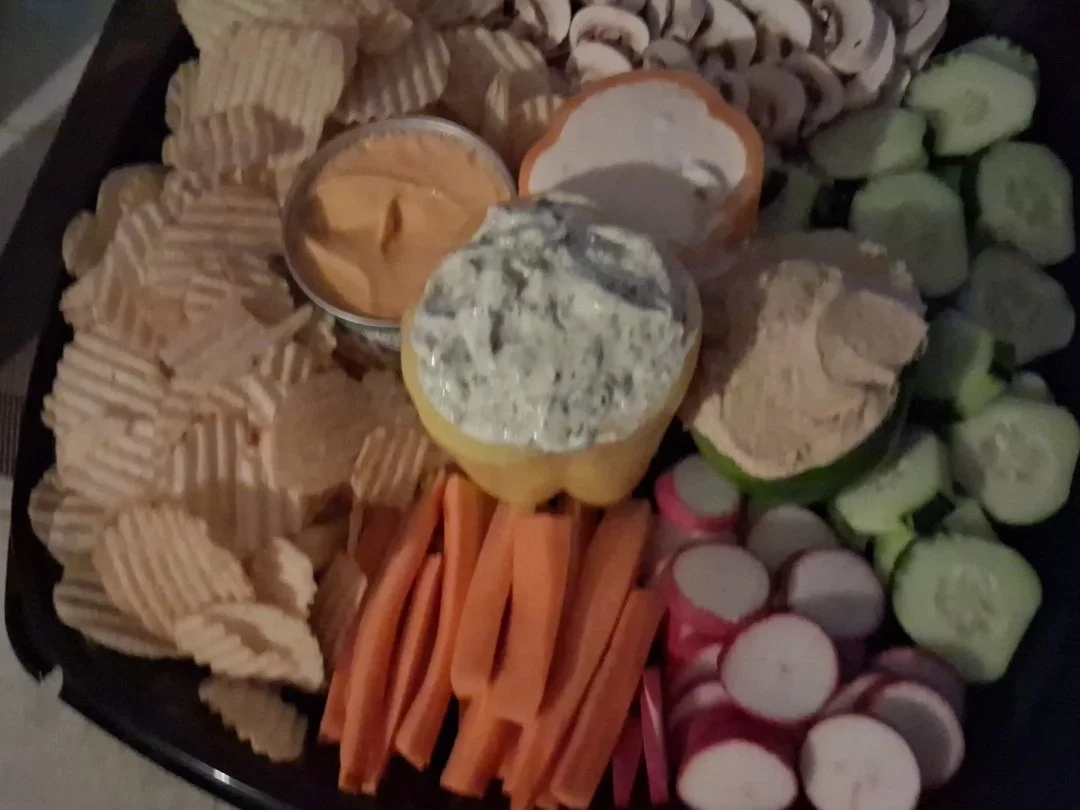 Assorted vegetable and snack platter with pita chips, carrot sticks, sliced cucumbers, radishes, and two dips.