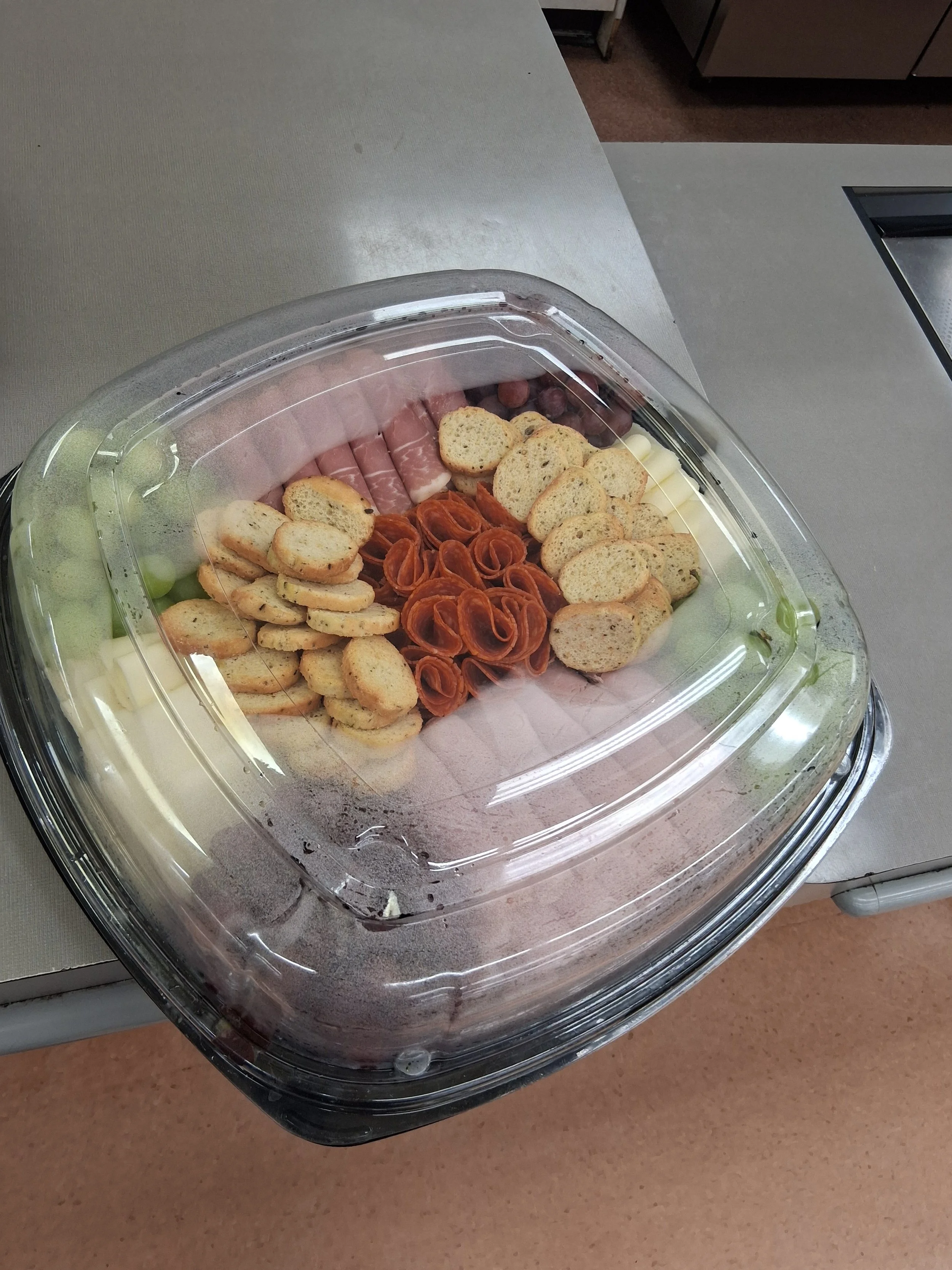 Clear plastic container with assorted cheeses, crackers, and cured meats on a countertop.