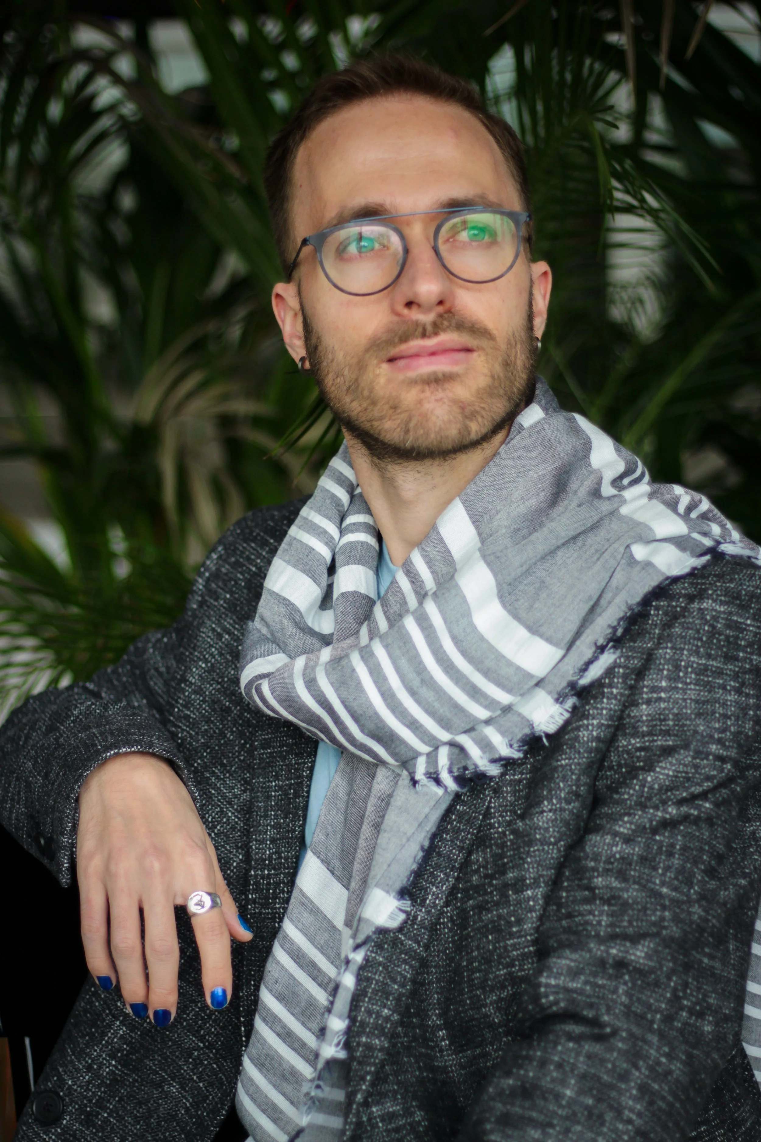 Portrait photograph of Andrin Albrecht, wearing a scarf, a dark blazer, and a light blue shirt, taken against a background of green plants. Credits to Olivia Fischer.