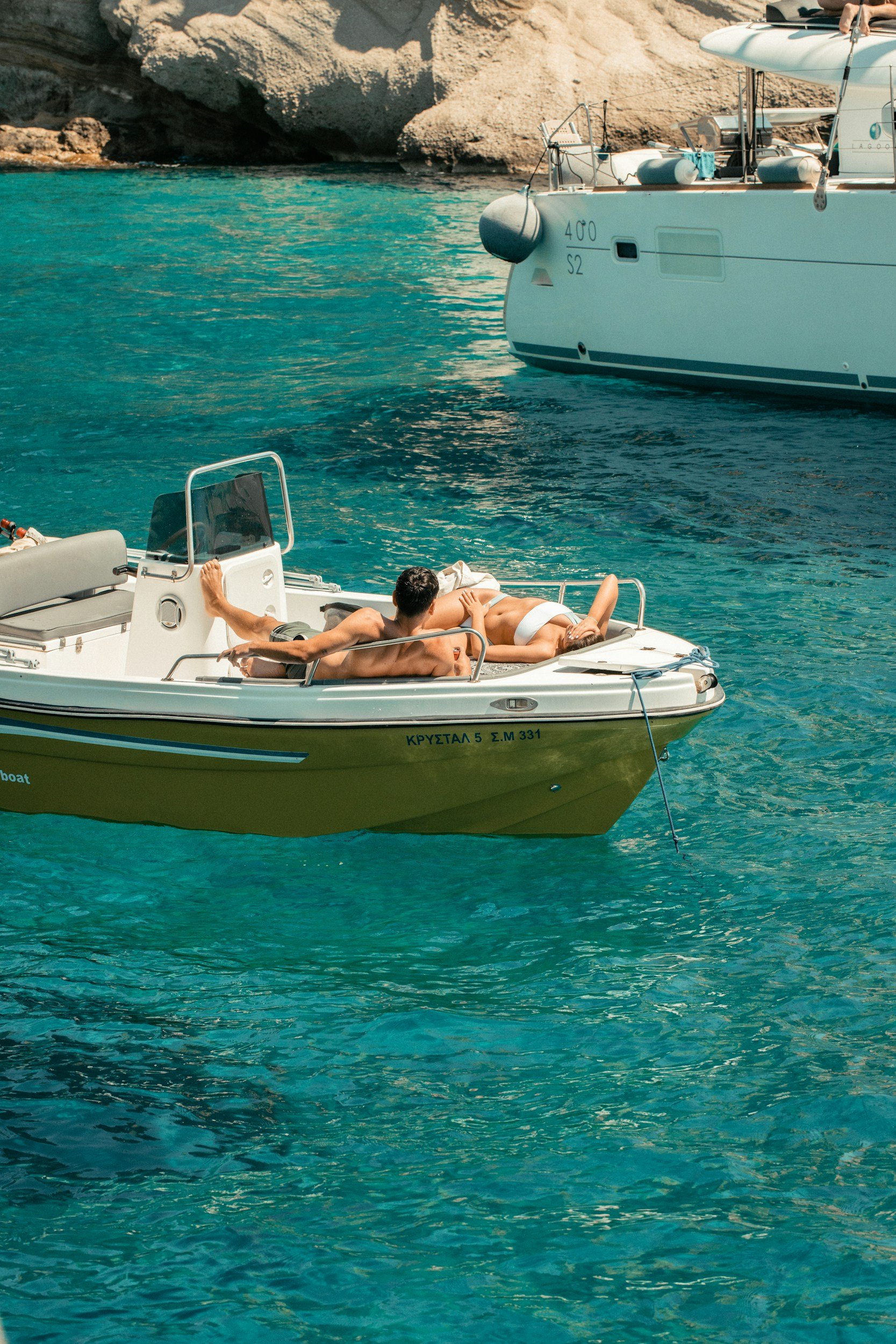 Two people relax on a small motorboat in clear blue water near a rocky shoreline, with another yacht anchored nearby.