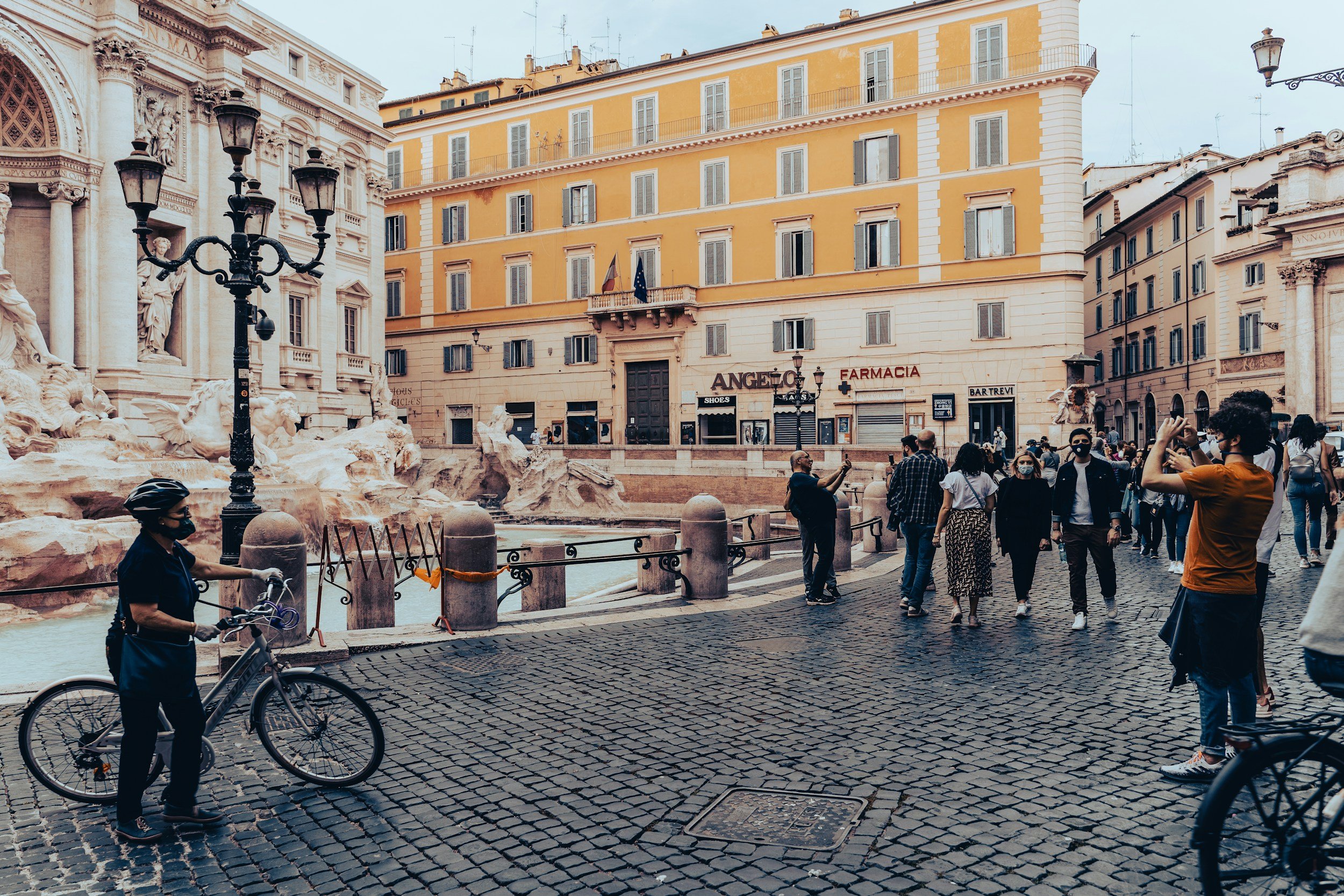 People walking and riding bikes on a cobblestone street near historic buildings, including a fountain and a church, under a cloudy sky in a busy urban square.