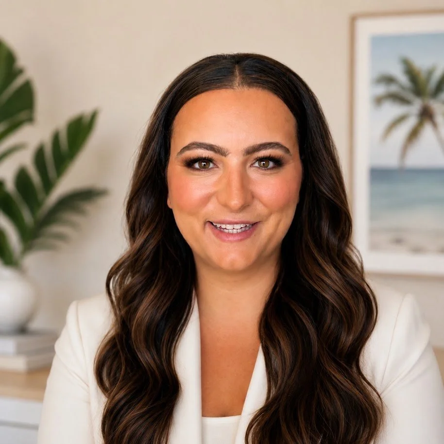 A woman with long dark hair and light makeup smiles at the camera in a bright room with a framed beach picture and a green plant in the background.