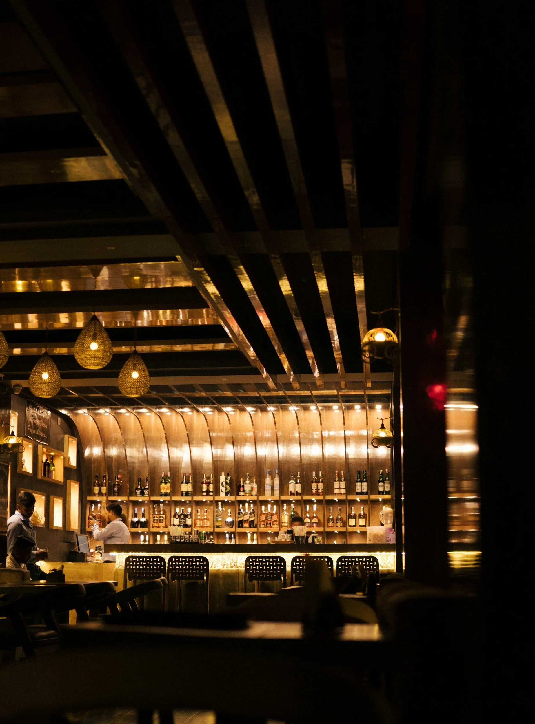 A dimly lit bar with shelves of liquor bottles, hanging pendant lights, and patrons seated at the bar and tables.