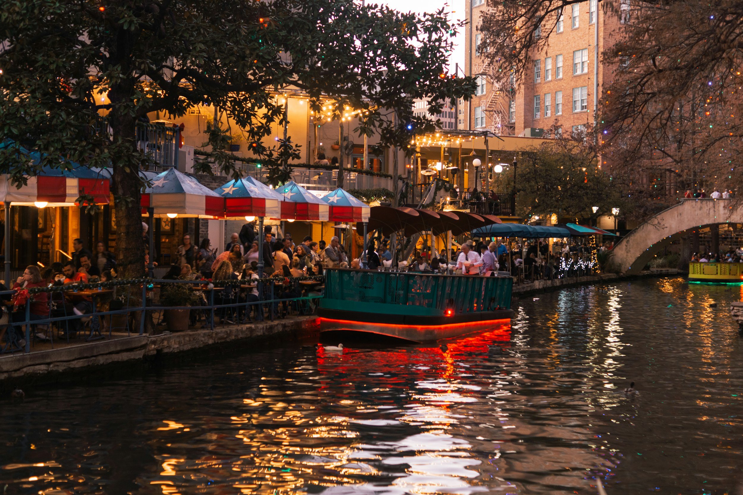 People gathered at a waterfront restaurant decorated with string lights and umbrellas featuring stars and stripes, with a boat moving through the water at dusk.