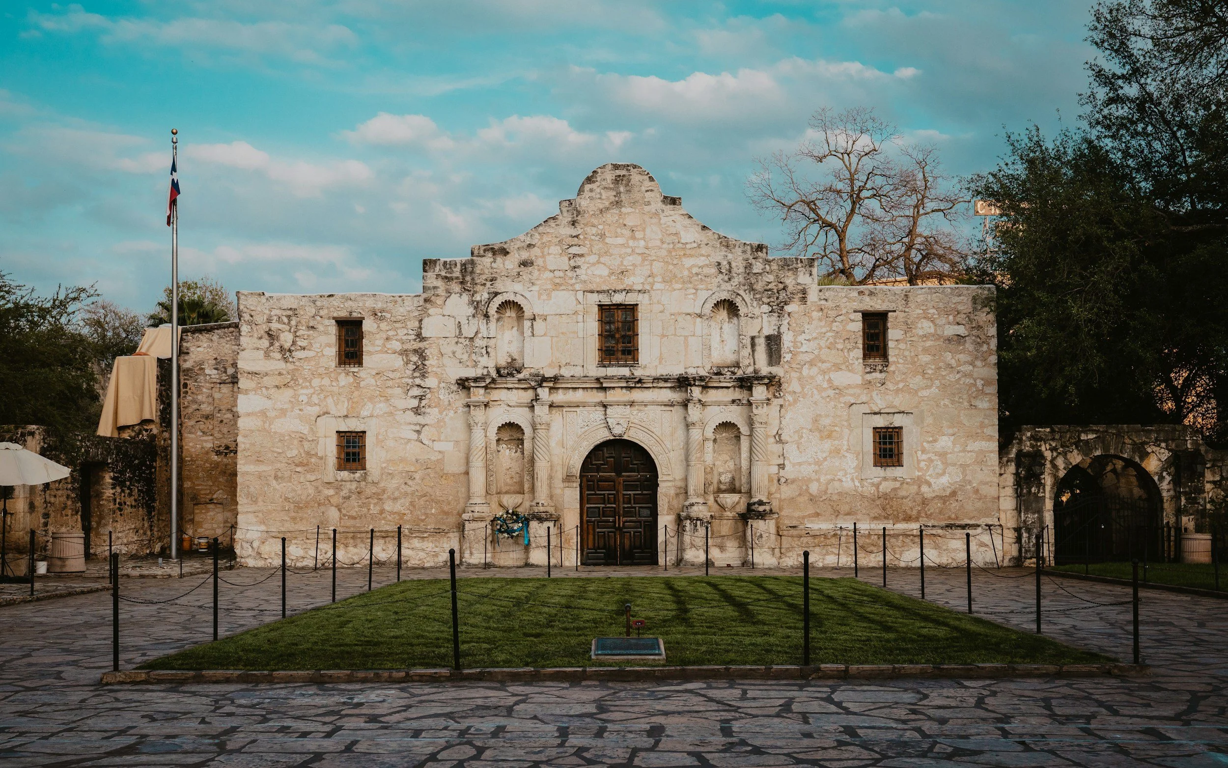 Front view of a historic stone building with an ornate entrance, arched windows, and a flagpole with the Texas flag. The building is surrounded by a paved courtyard, fencing, and trees, with a partly cloudy sky in the background.