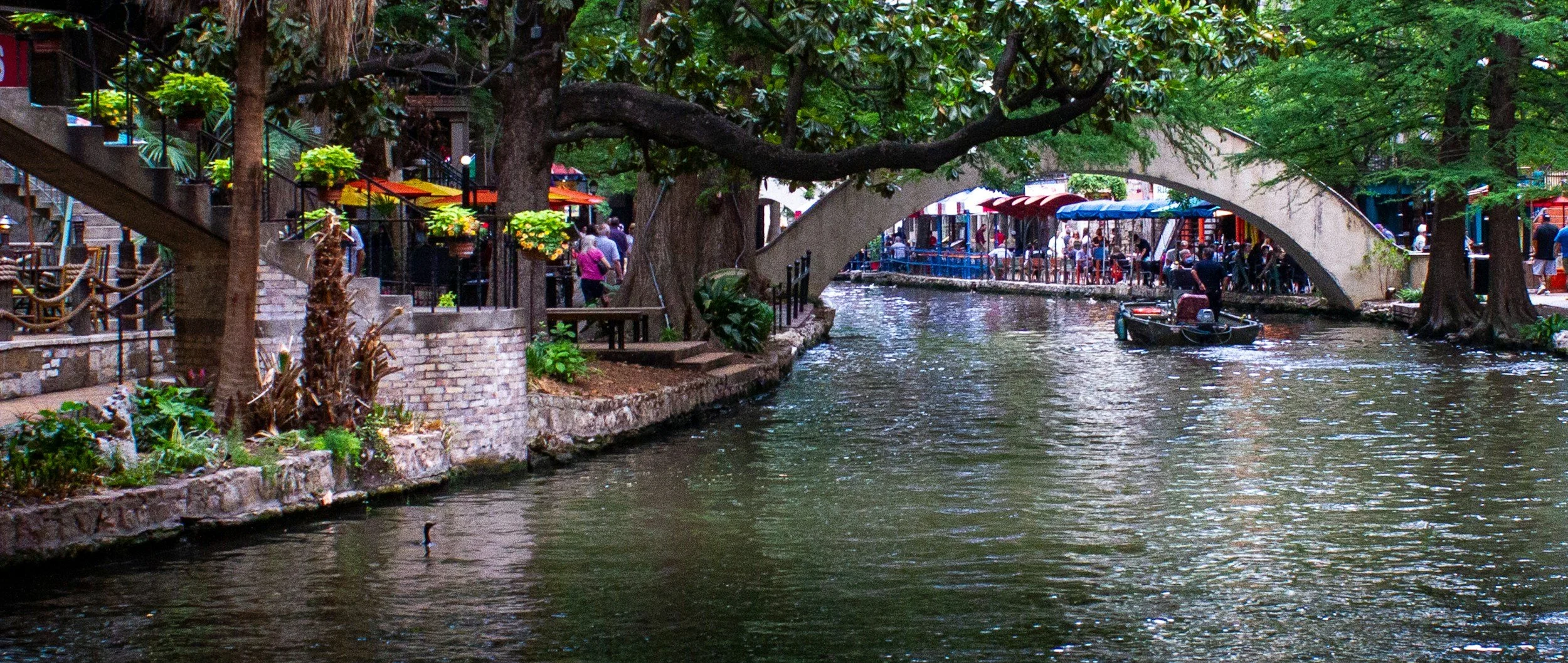 A lively downtown area along a waterway with a stone bridge, trees, and shops with colorful awnings, and a boat on the water.