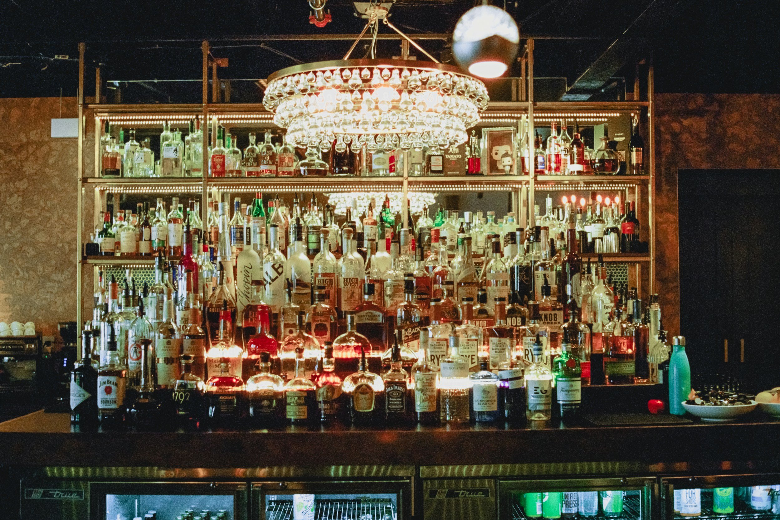 A bar with a large chandelier made of glasses hanging in the center. The bar is stocked with various bottles of alcohol, and there are glasses and barware displayed on the shelves. The background is dark, and the lighting highlights the bottles and t