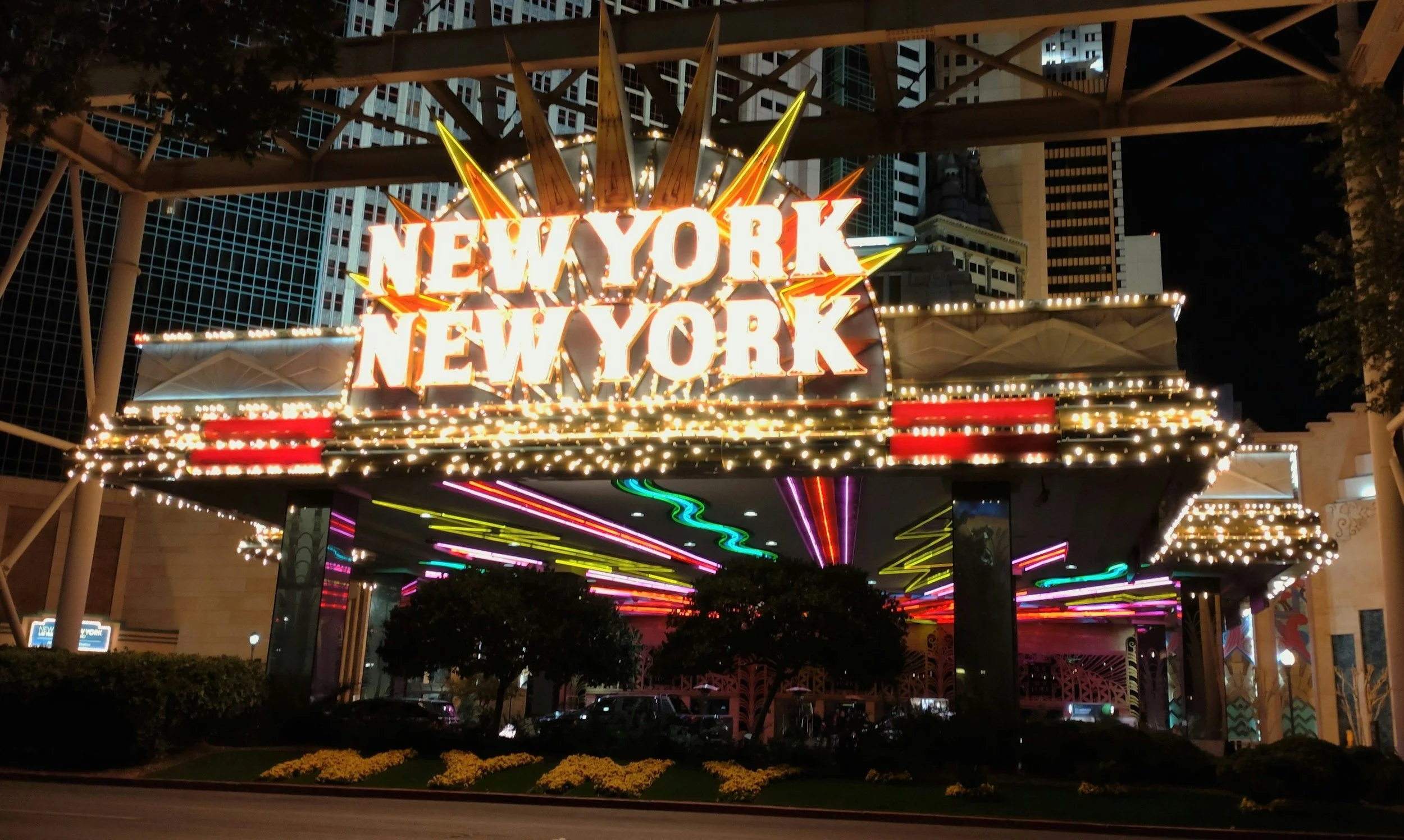 Nighttime view of a brightly lit New York New York sign at an amusement park, with colorful light streaks on a ride below and tall buildings in the background.