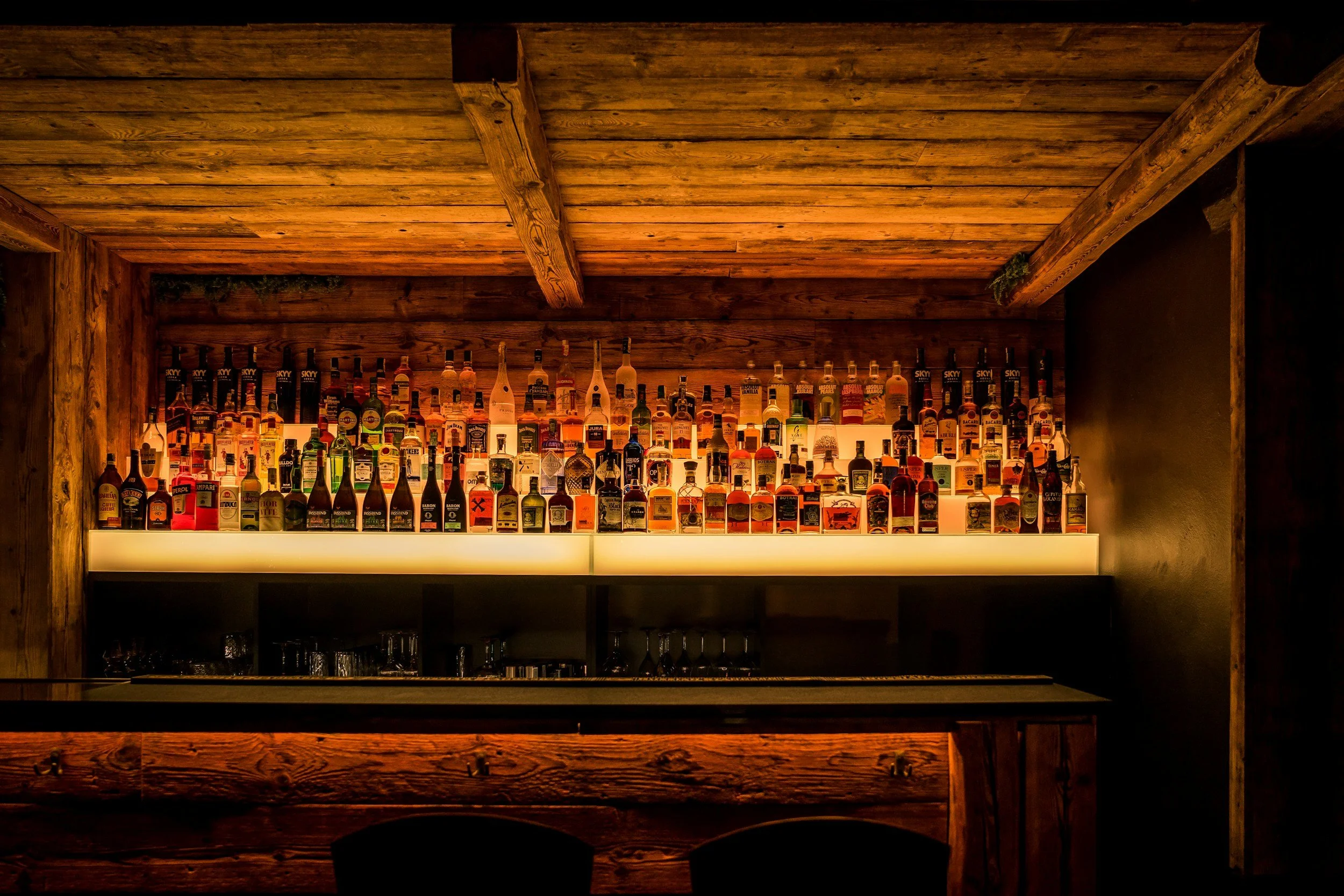 A bar shelf displaying various bottles of alcohol under warm lighting, with a wooden ceiling and walls.