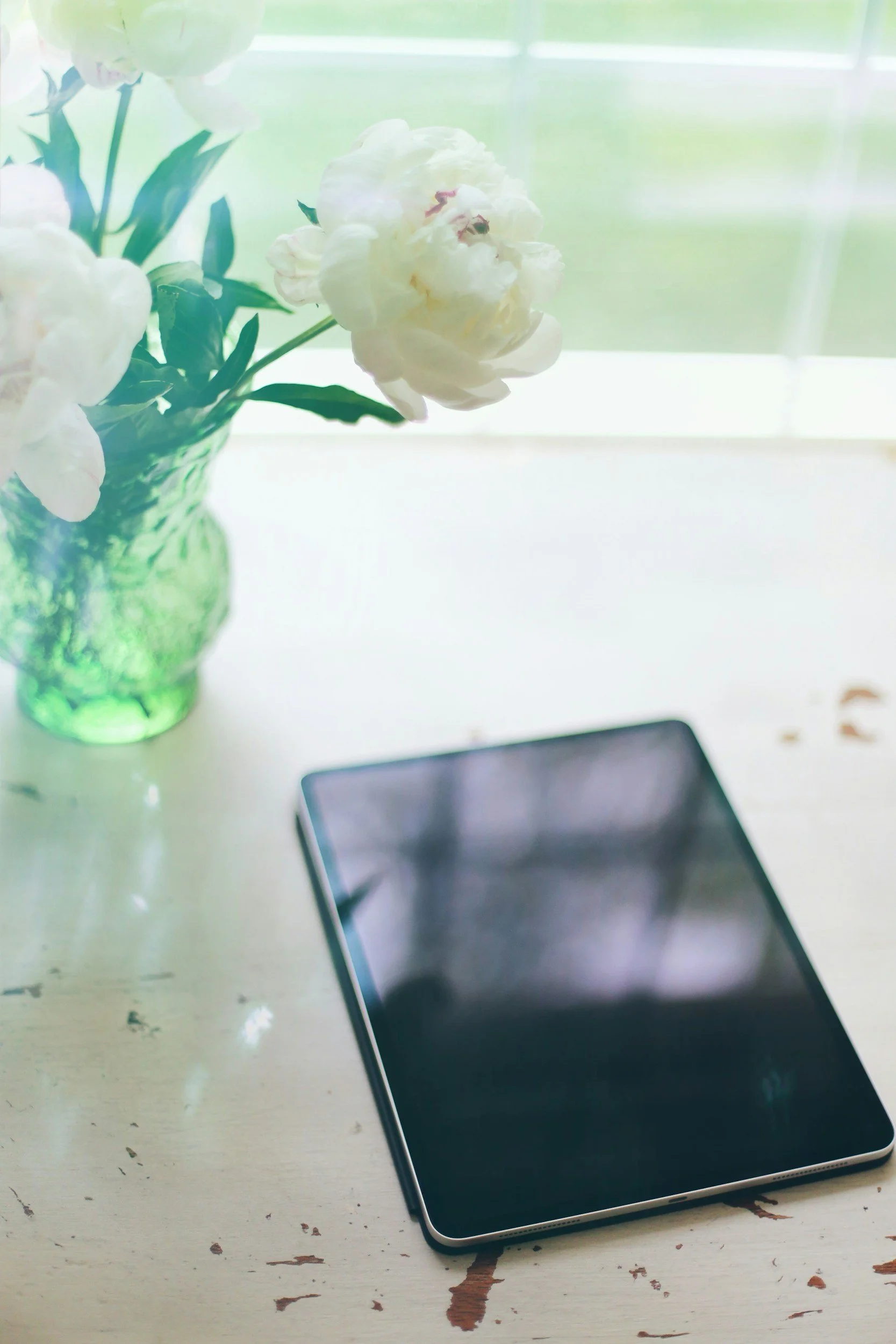 A white table with a black smartphone and a green glass vase with white peonies. The background shows a window with green scenery outside.