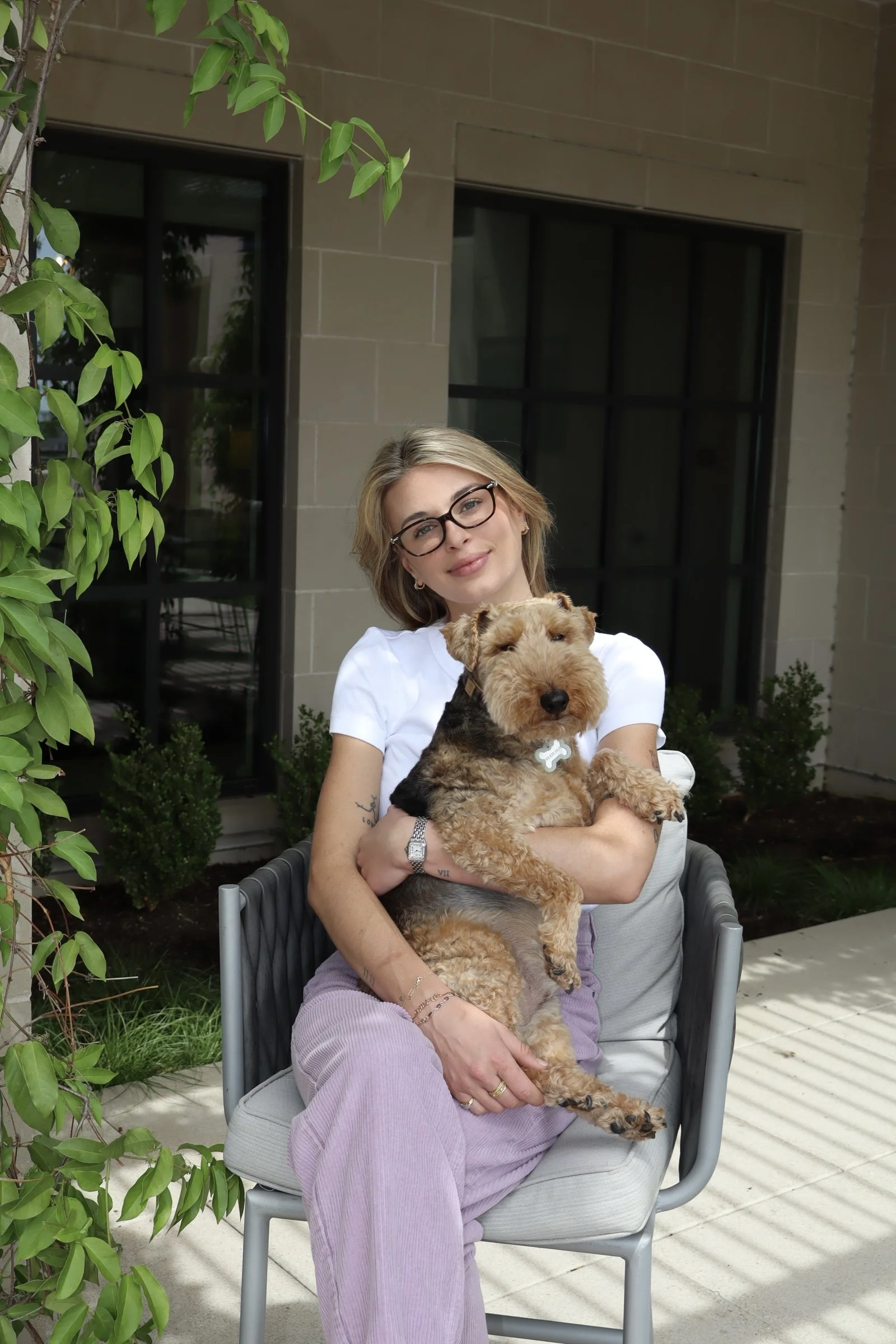A woman with blonde hair and glasses holding a tan and black dog in her lap, sitting outside near a beige building with large windows and green foliage.