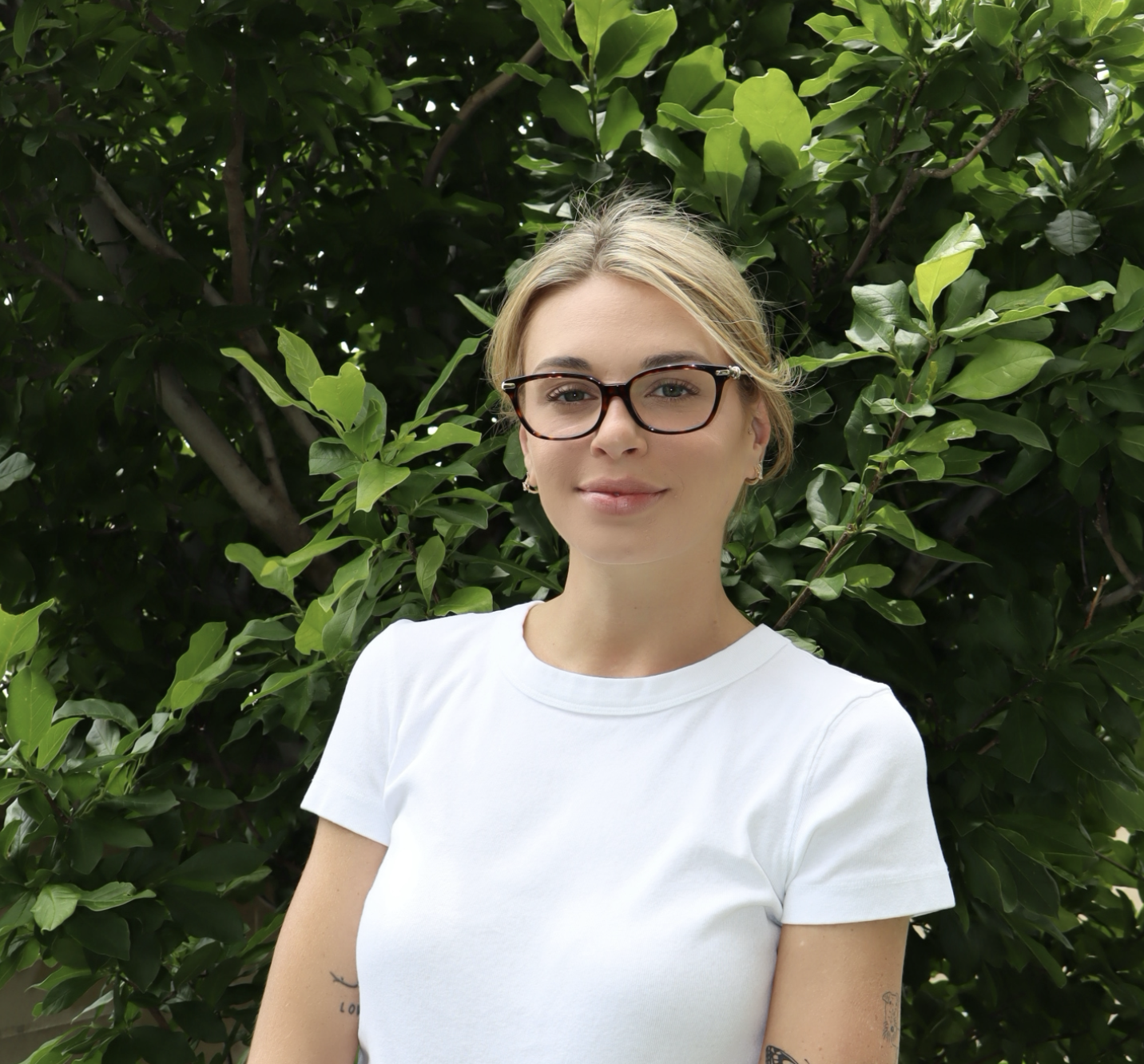 A woman with glasses and blonde hair wearing a white t-shirt stands outdoors in front of green leafy bushes, smiling gently.