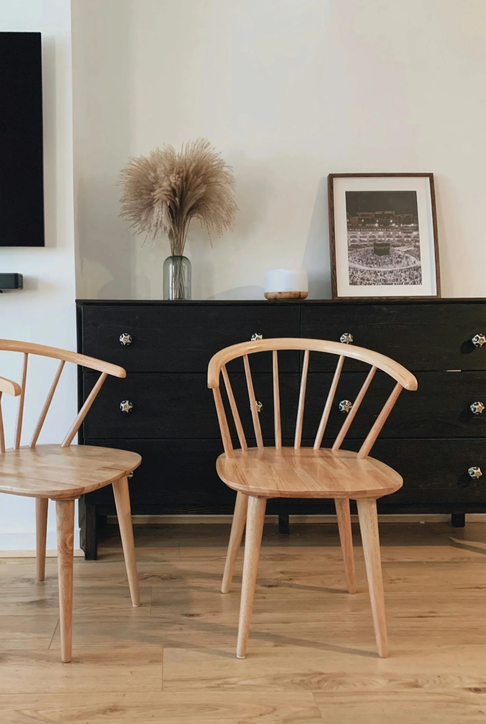 Interior of a dining area with two wooden chairs in the foreground, a black dresser with a framed black-and-white photo, a vase with pampas grass, and a small white object on top of the dresser, against a plain light-colored wall.