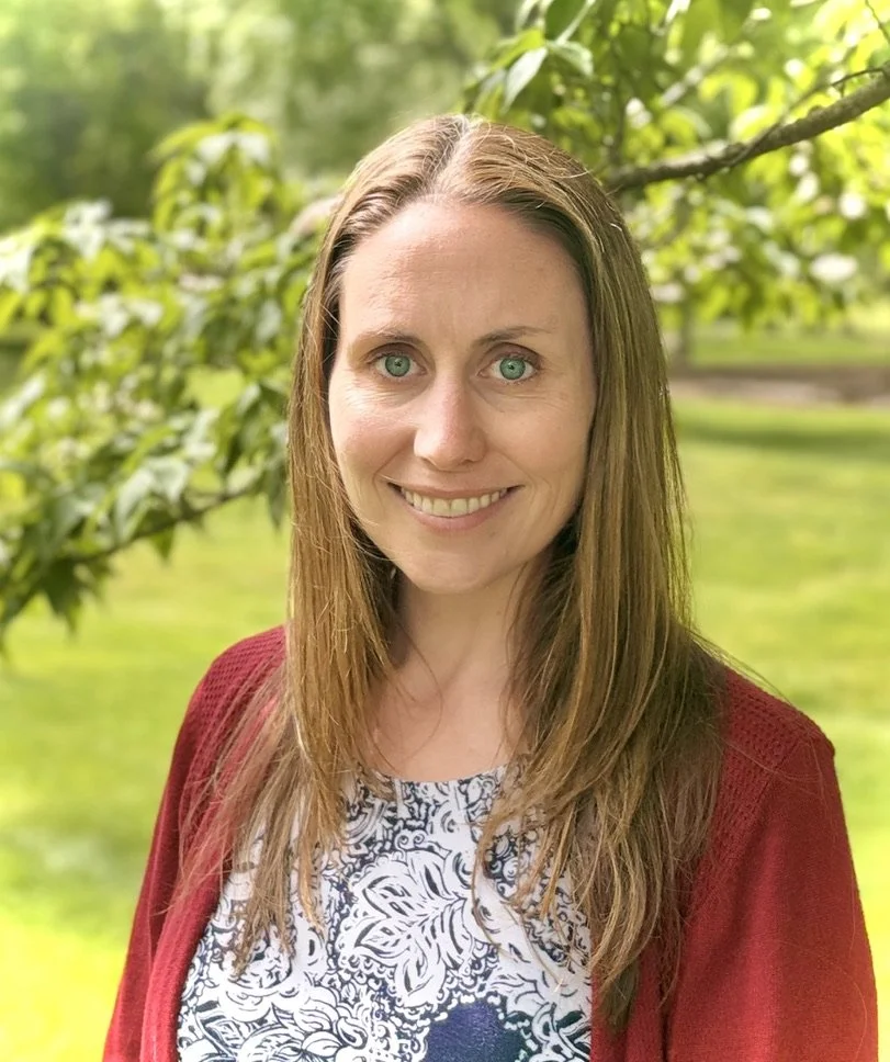 A woman with long, light brown hair and blue eyes smiling outdoors with green trees and grass in the background.